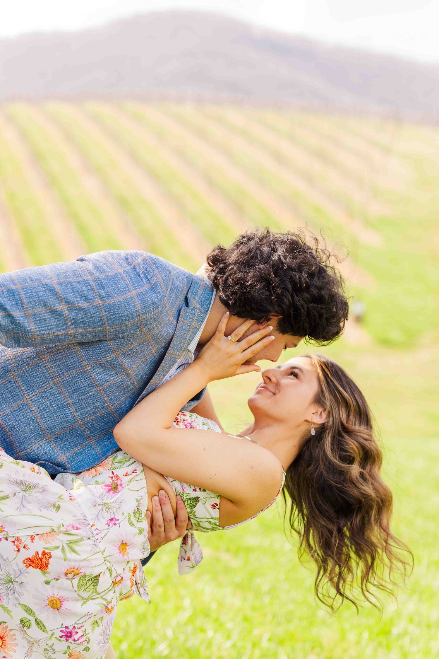 Man leaning down to kiss woman as she arches back gracefully in front of a scenic vineyard landscape.