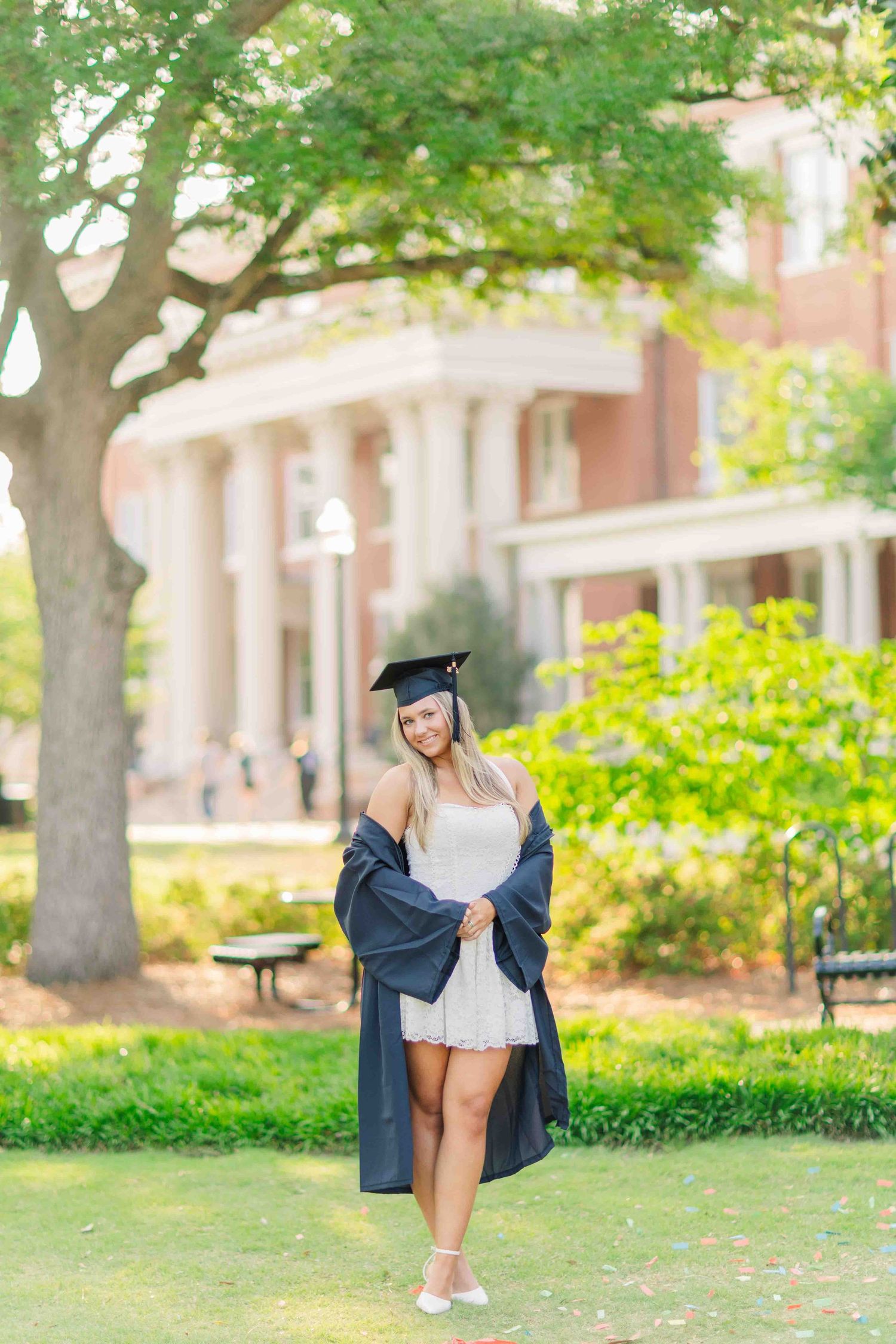 Smiling graduate in cap and gown over white dress poses outdoors on college campus green lawn.