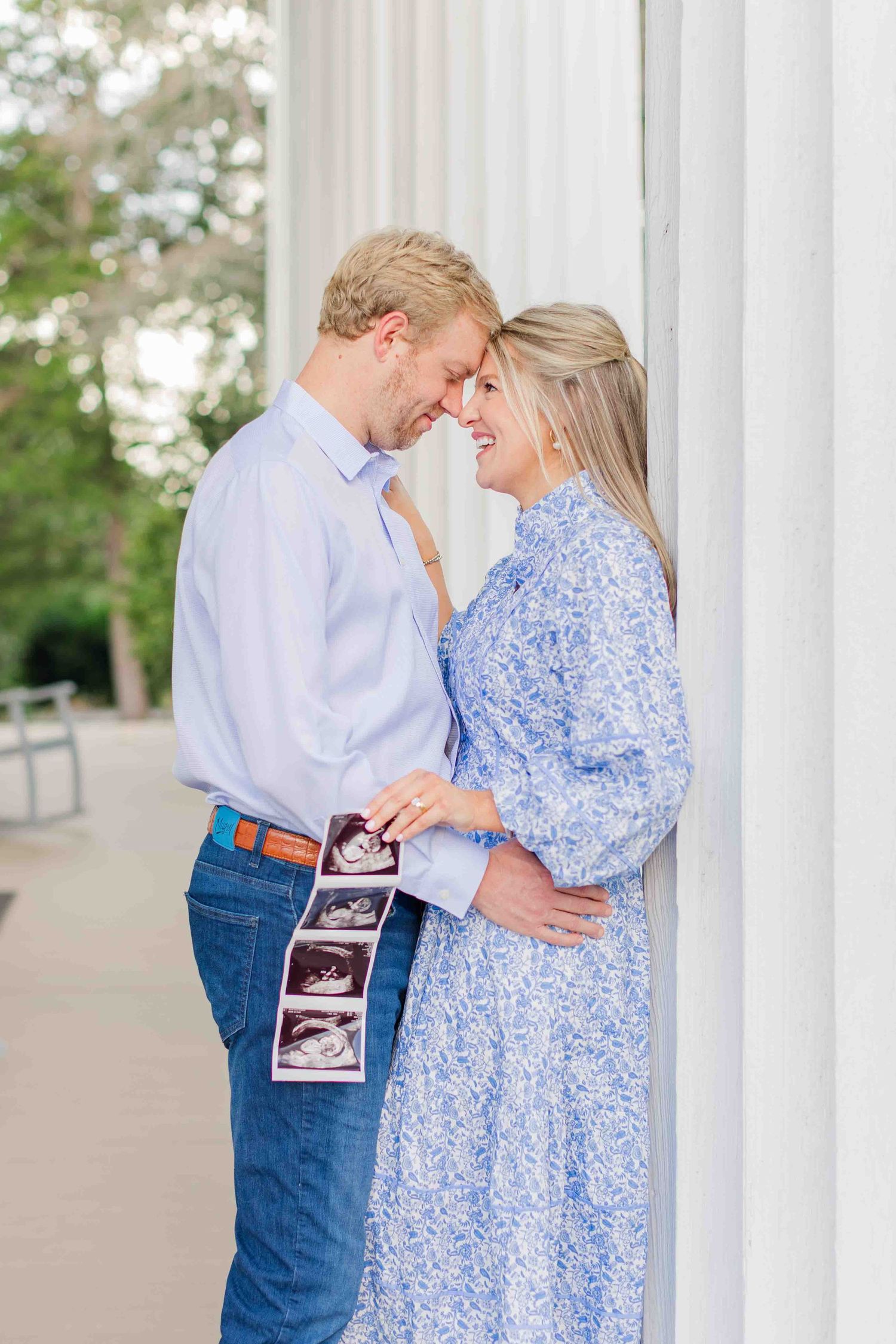 Couple sharing a tender moment holding ultrasound photos, announcing pregnancy near white columns outdoors.