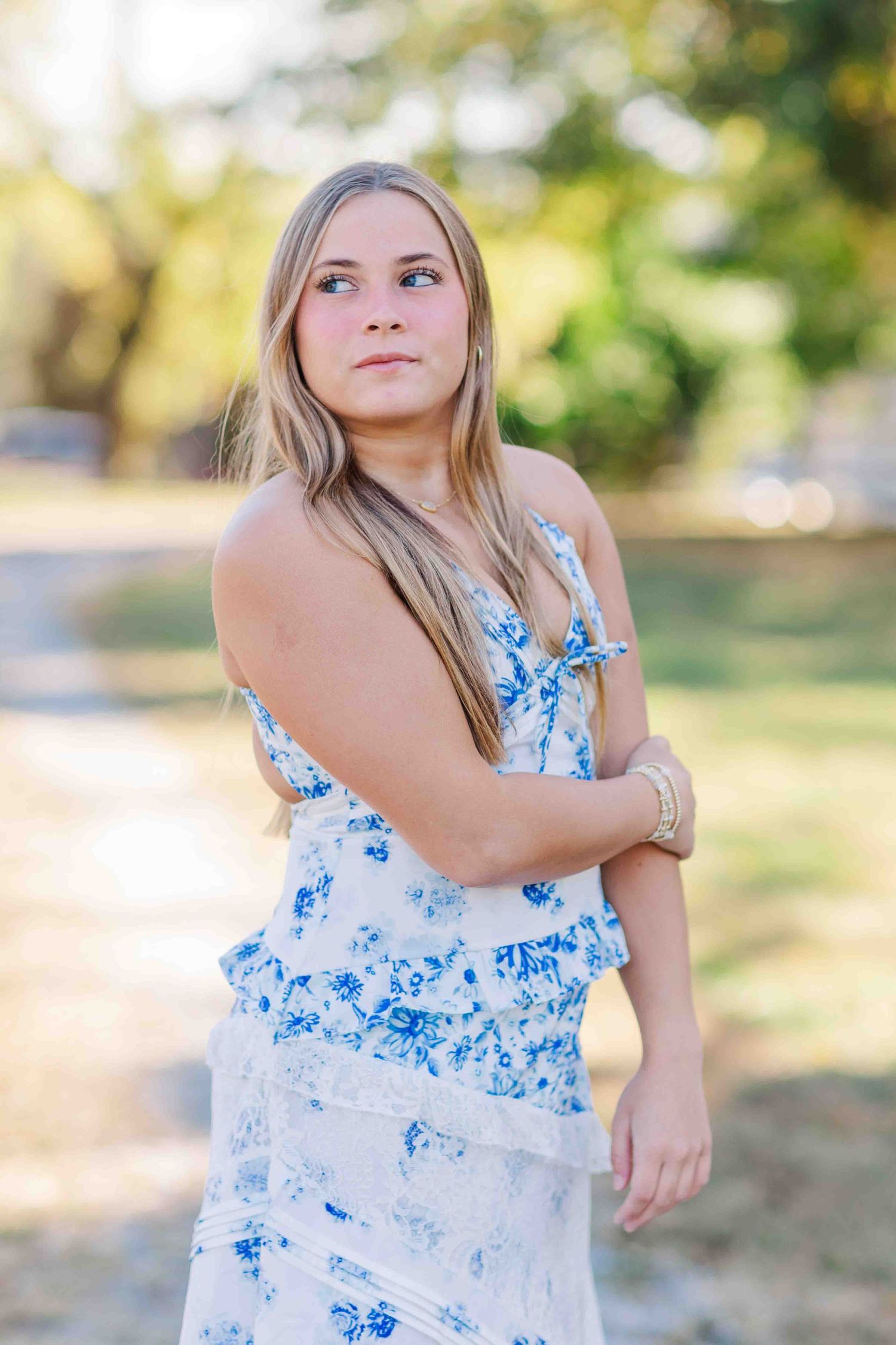 Blonde woman in blue and white floral dress posing outdoors in a sunlit park setting.