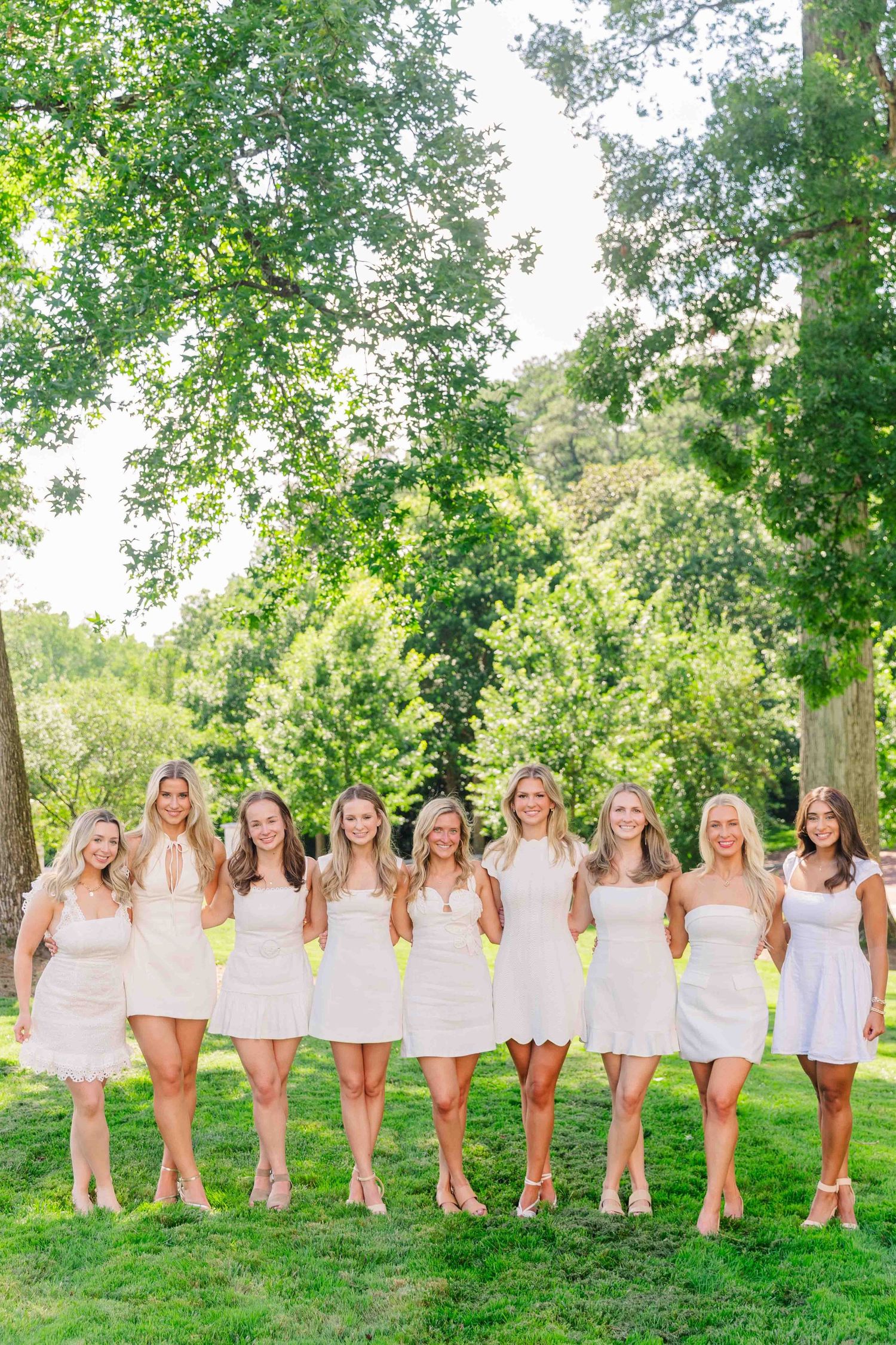 Nine women in white dresses posing together outdoors on green grass surrounded by trees.