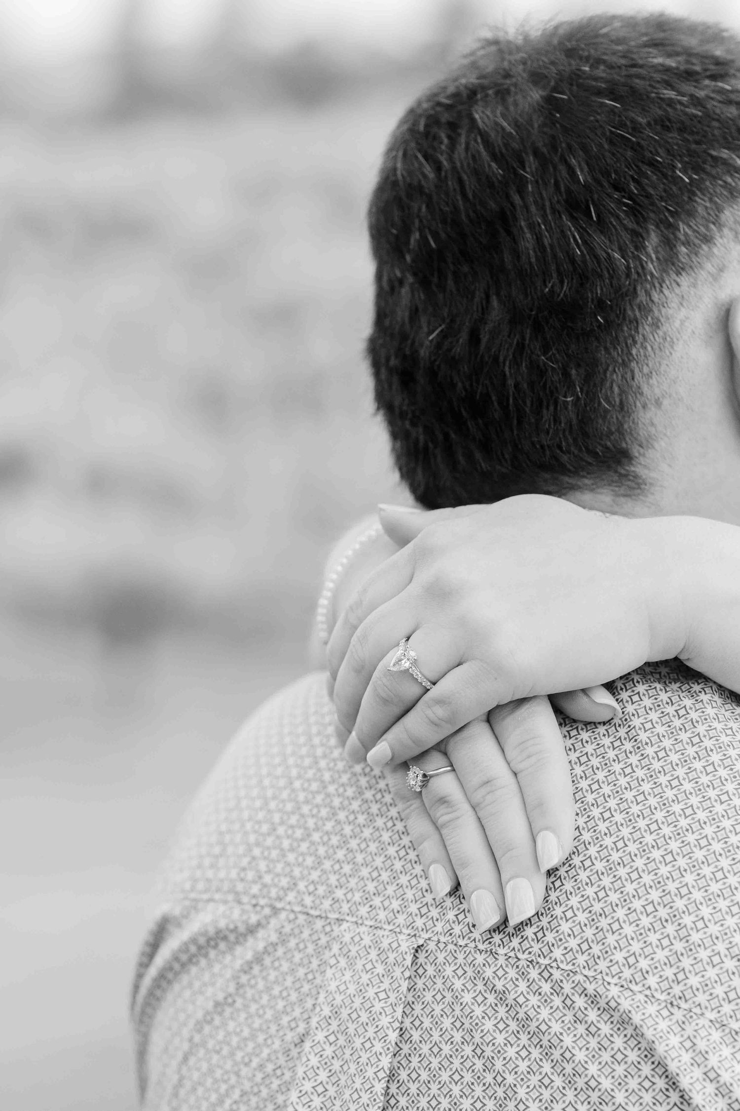 Black and white close-up of woman's hand with diamond ring resting on man's shoulder in patterned shirt.