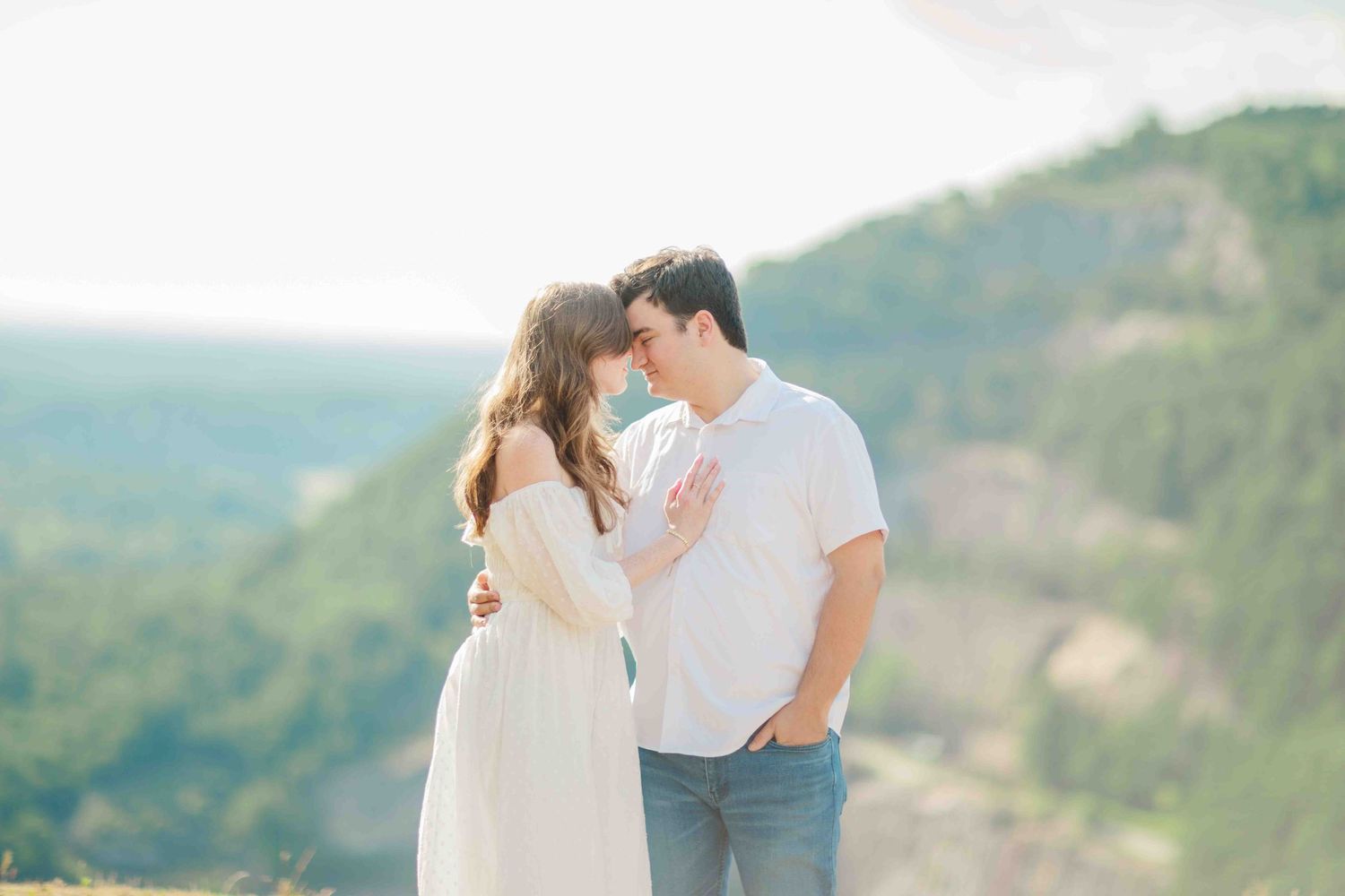 Couple embracing and touching foreheads on a scenic mountain overlook, dressed in white and jeans.