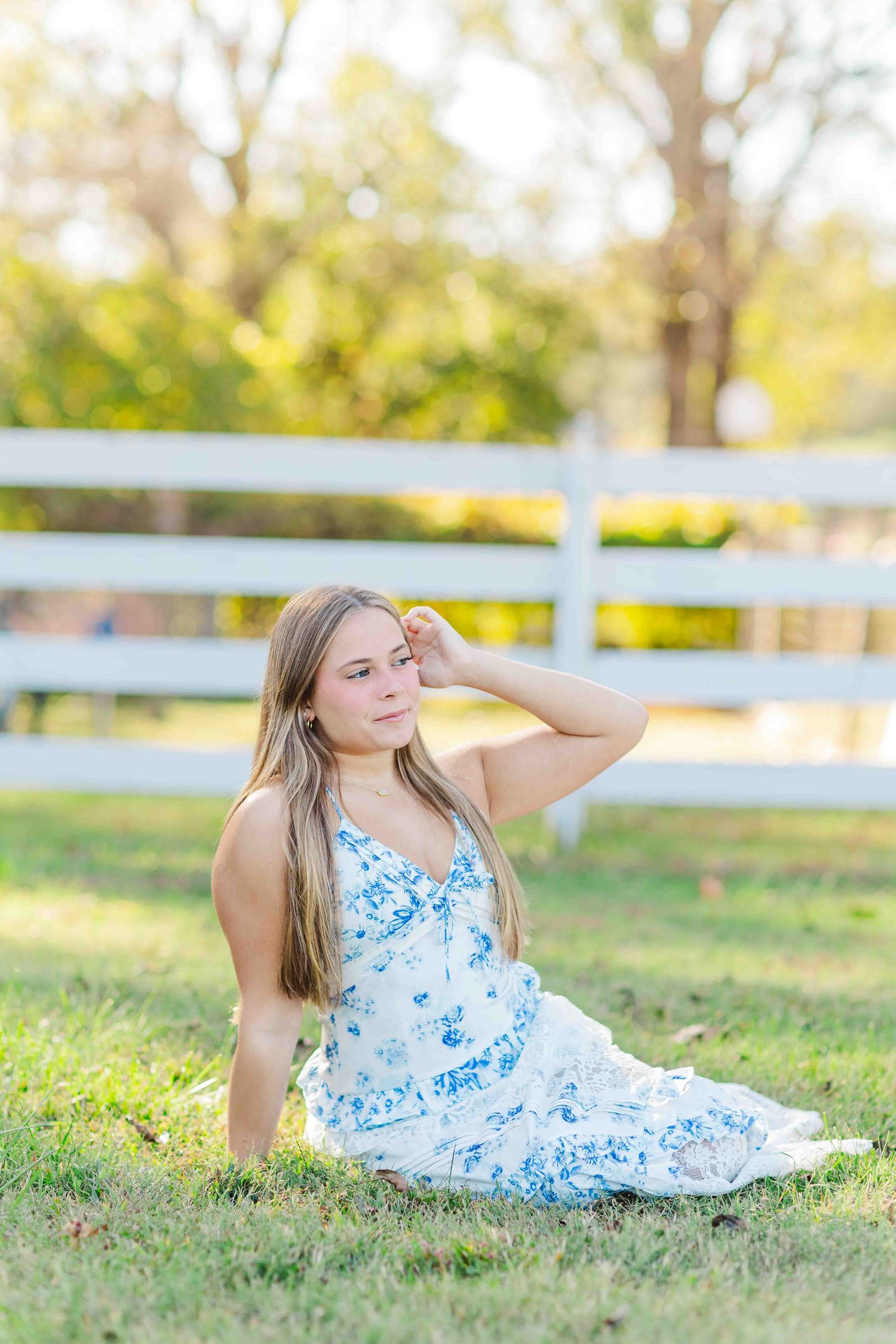 Blonde woman in white floral dress sitting on grass by white fence, hand raised to her hair.