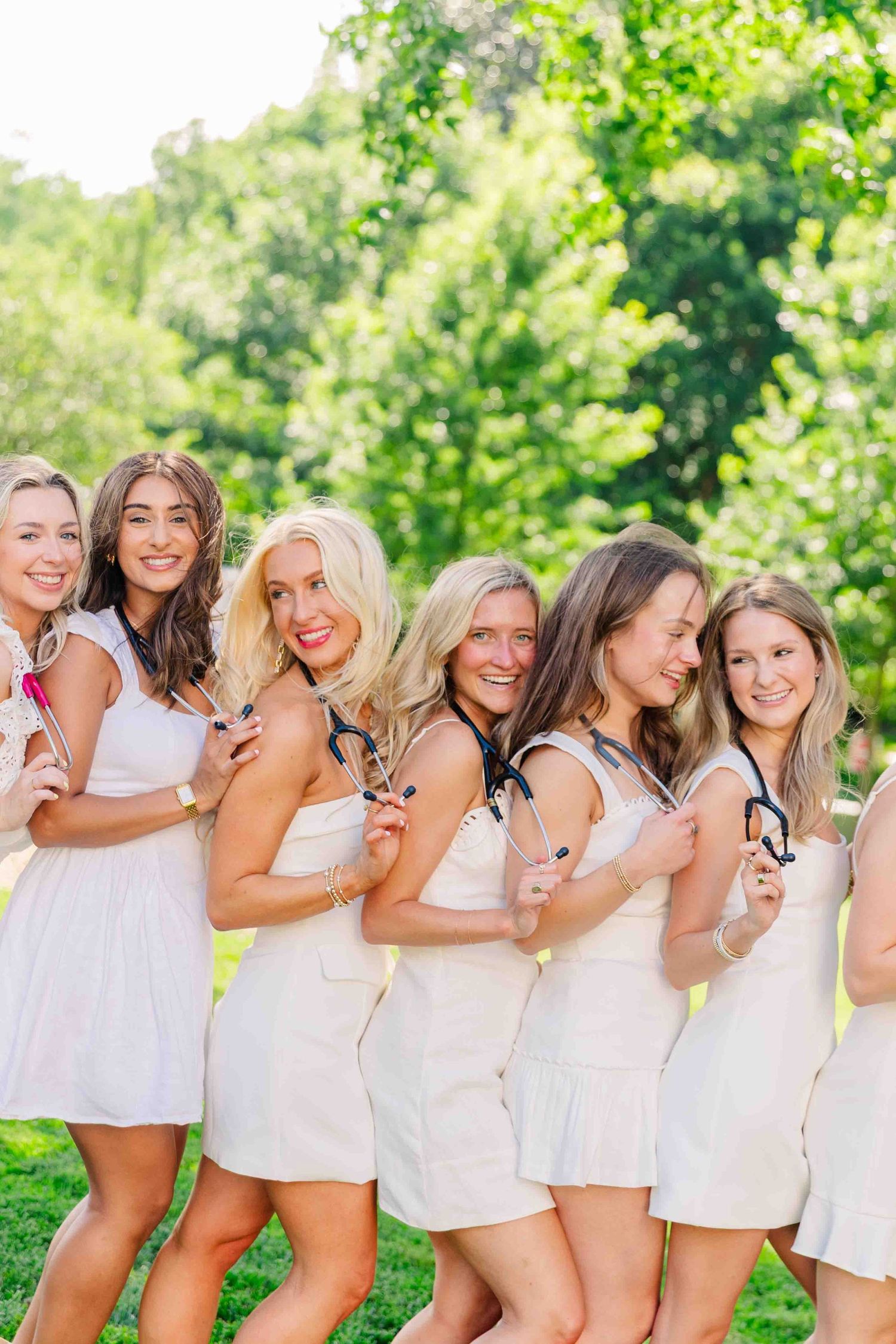 Six women in white dresses posing with stethoscopes in lush green outdoor setting.