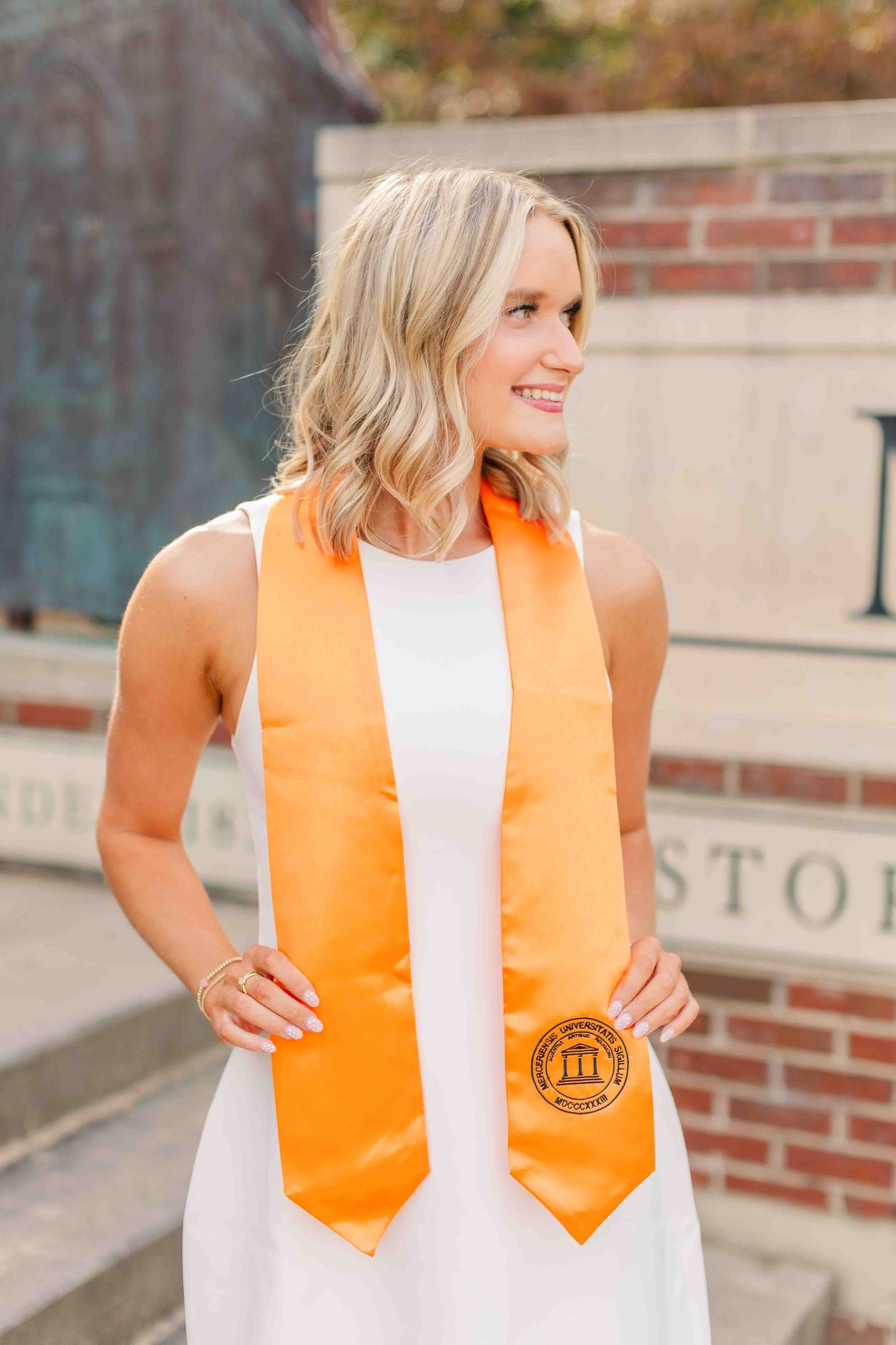 Smiling blonde woman in white dress wearing an orange university graduation stole outdoors.