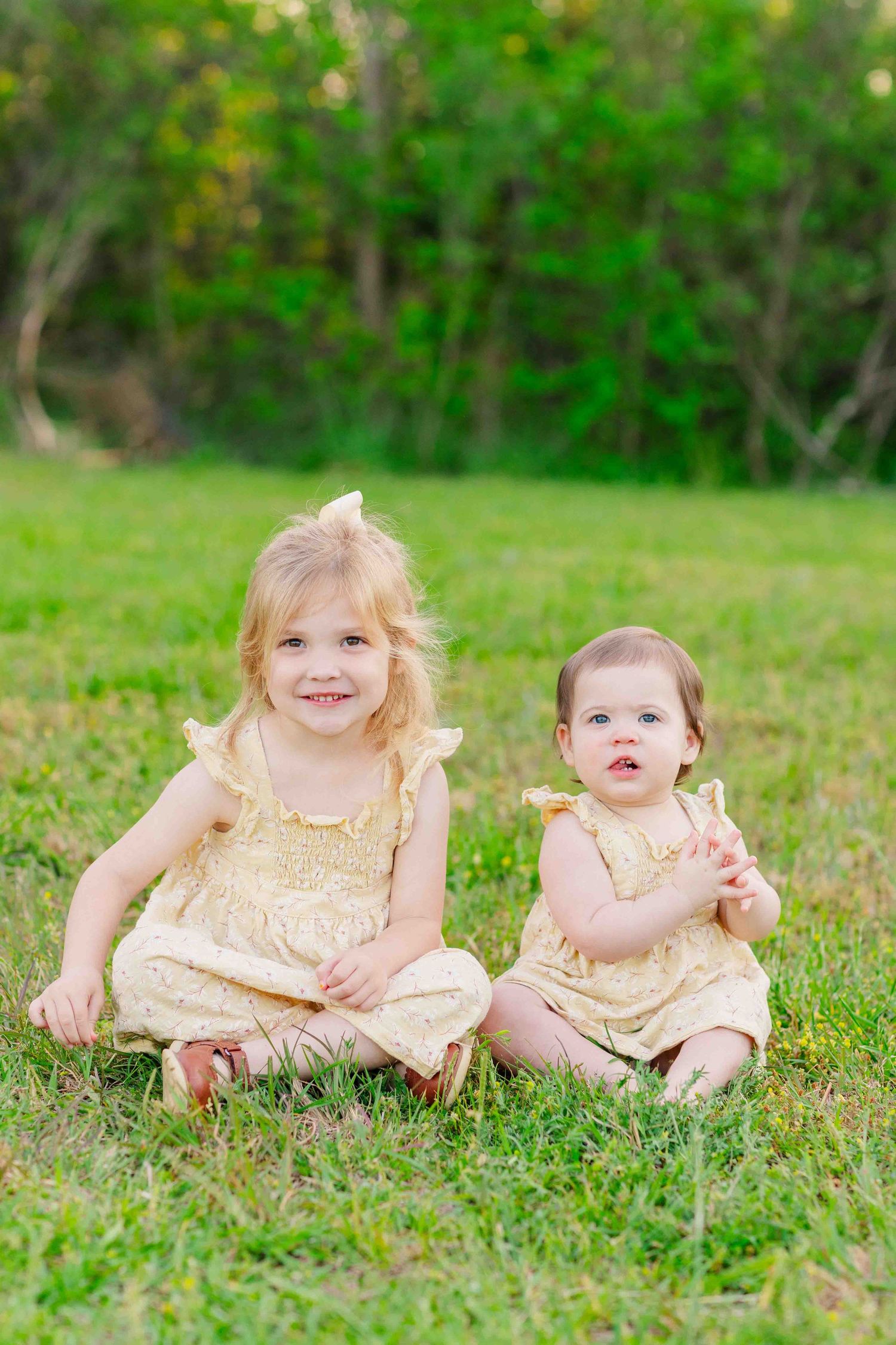 Two adorable sisters in matching yellow outfits sitting together smiling on a green grass lawn.