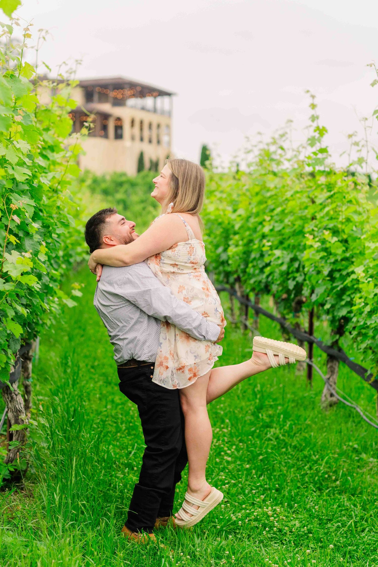 Man lifting woman joyfully in his arms between green vineyard rows with winery building behind them.