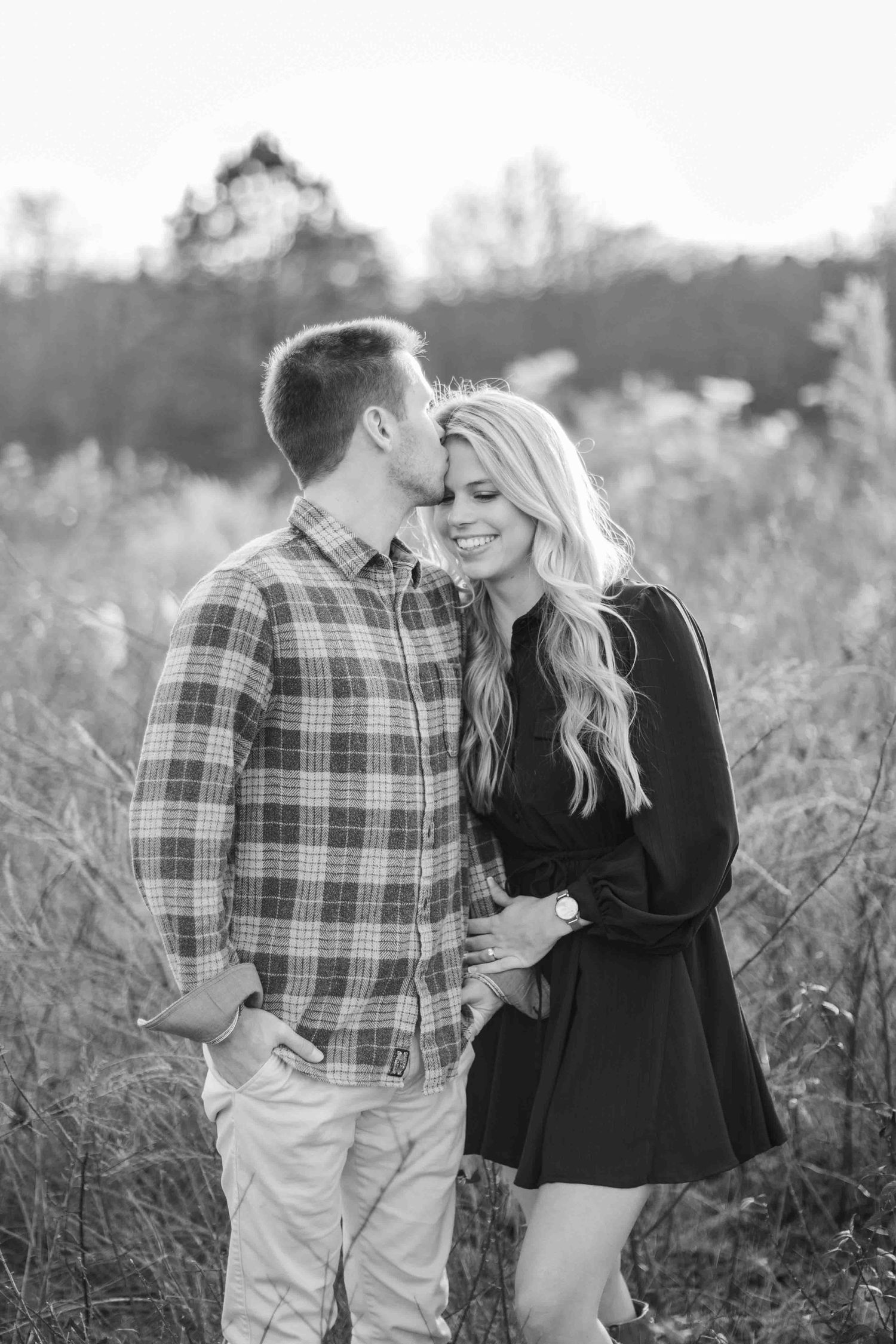 Black and white photo of couple in field, man kissing woman's cheek as she smiles warmly.