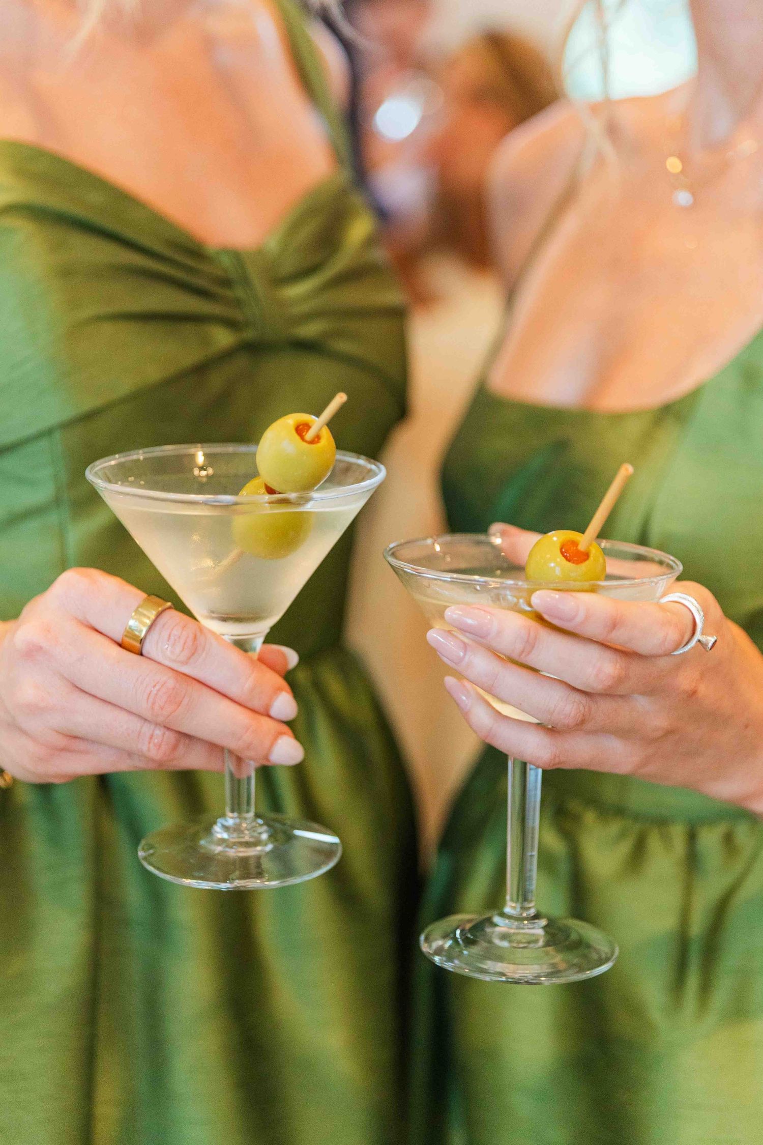 Two women in green satin dresses holding dirty martini cocktails garnished with olives on toothpicks.