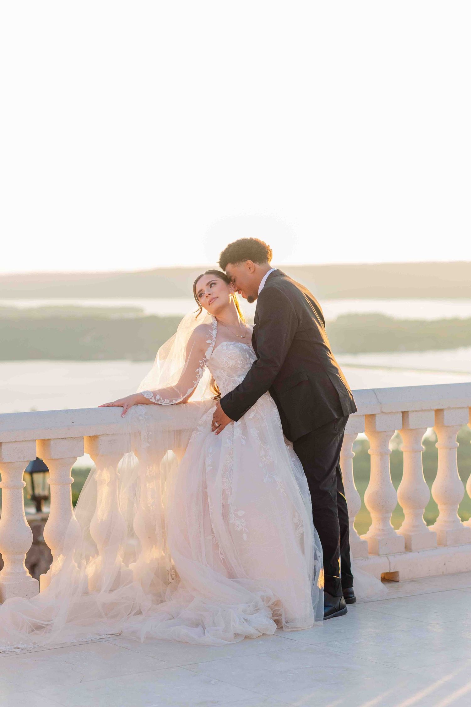 Bride and groom share a romantic moment on a sunlit balcony with scenic water views on their wedding day.