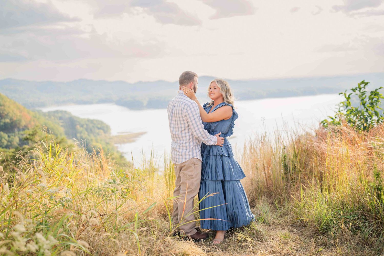 Engaged couple embracing on a hilltop with a serene lake and lush green mountains in the background.