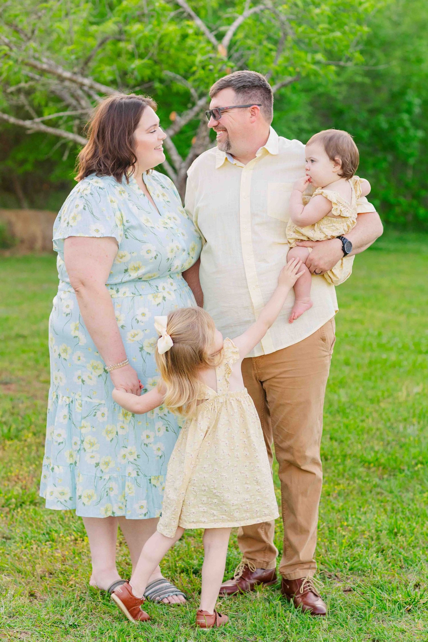 Happy family of four with two young daughters holding hands and smiling in a green outdoor setting.