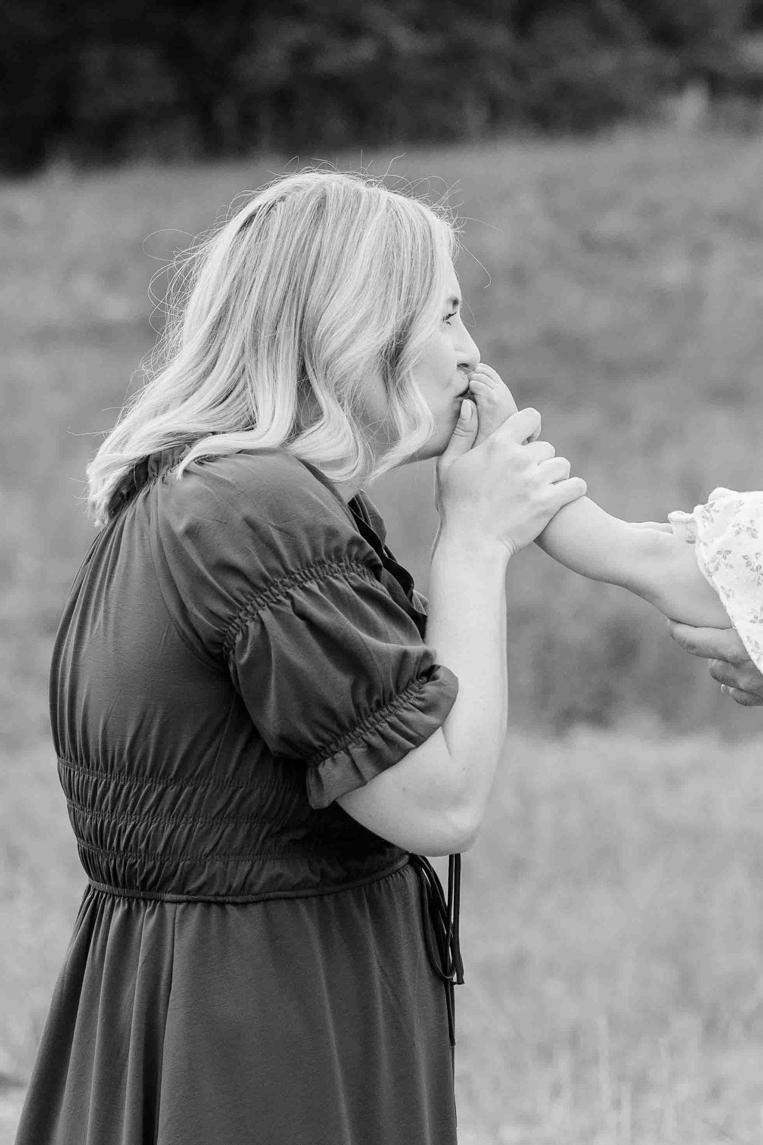 Black and white photo of a mother tenderly kissing her childs hand in an outdoor field.