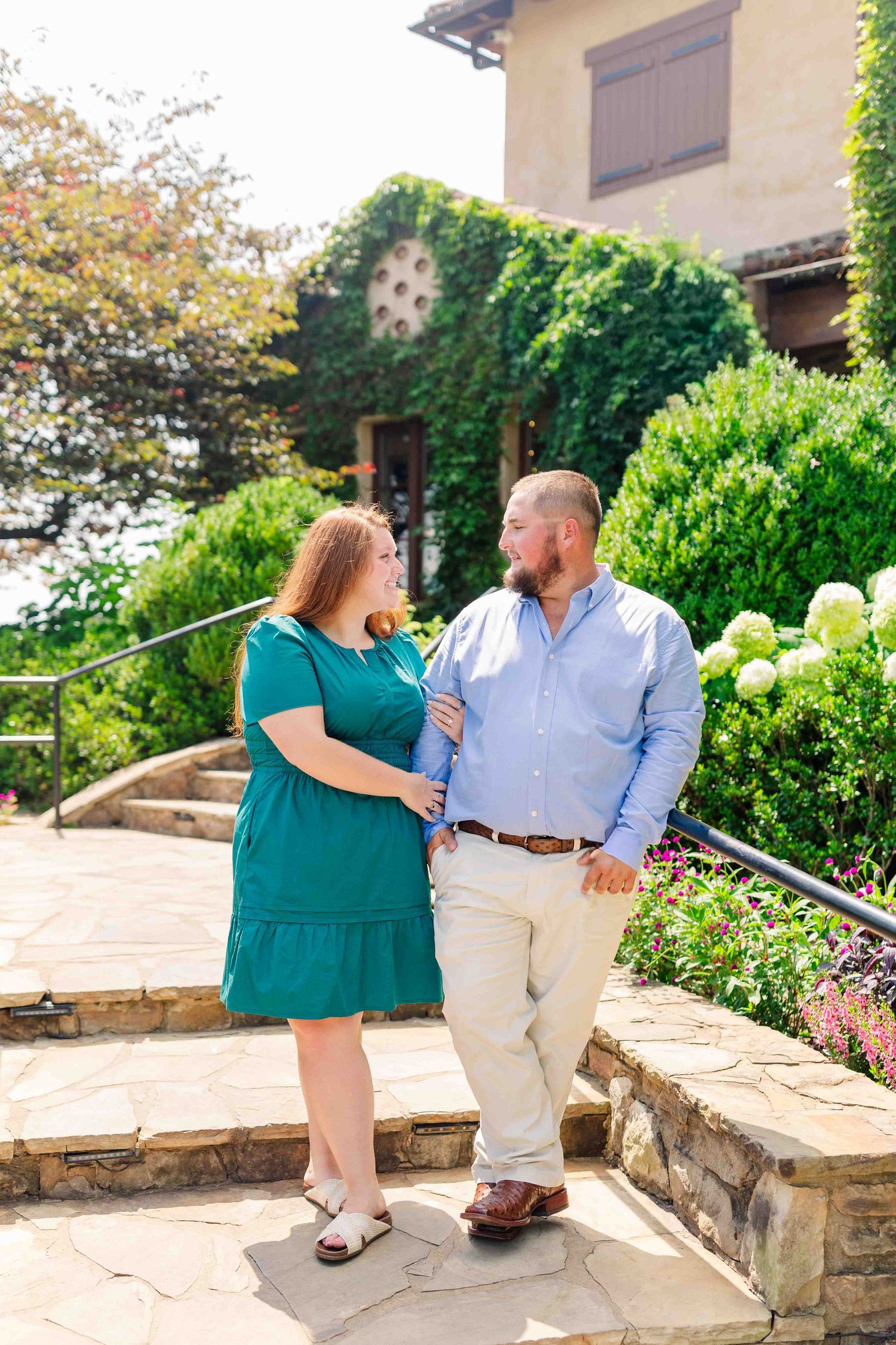 Engaged couple smiling at each other on stone steps outside ivy-covered winery surrounded by lush gardens.