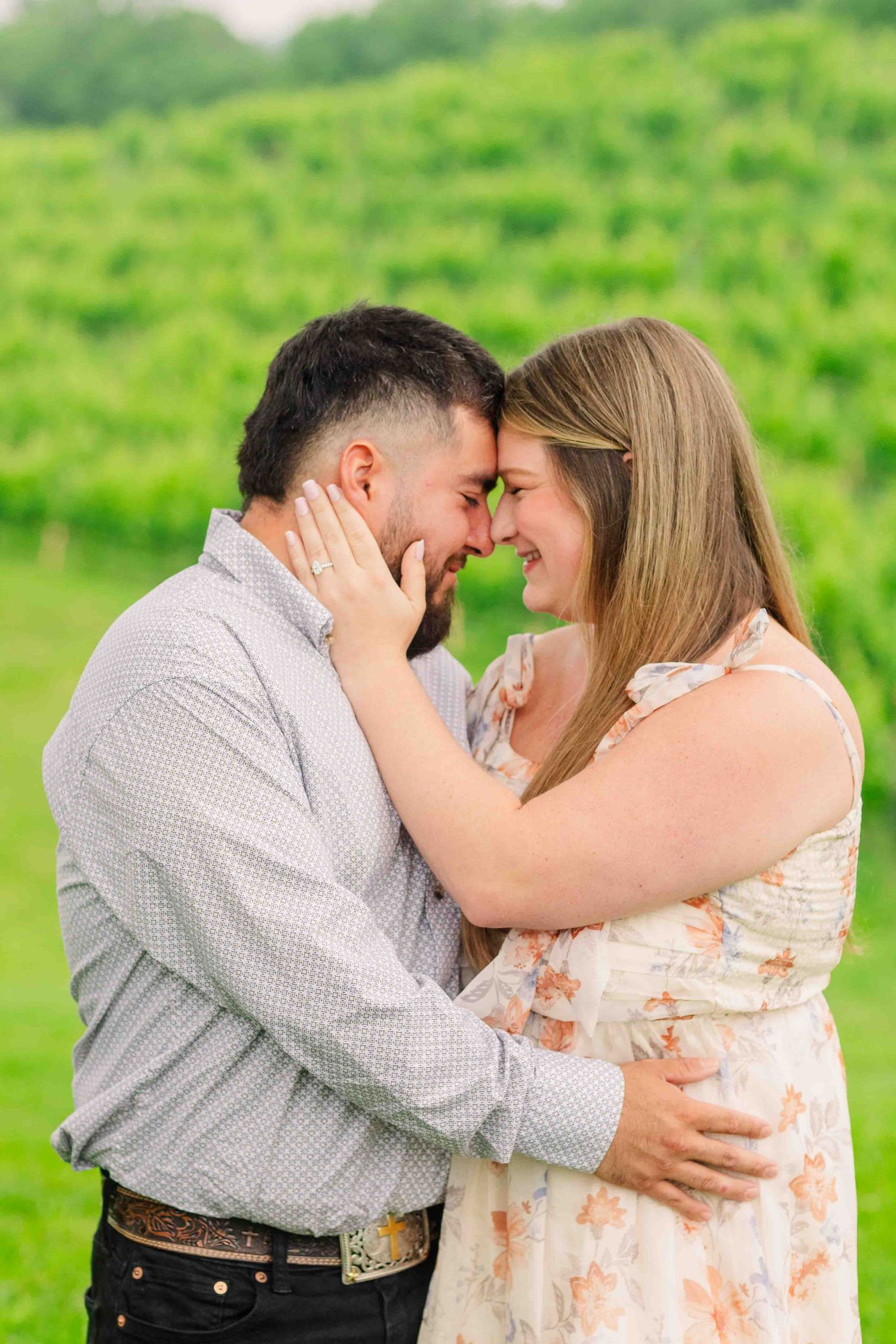 Romantic couple touching foreheads and embracing in front of vibrant green vineyard on overcast day.