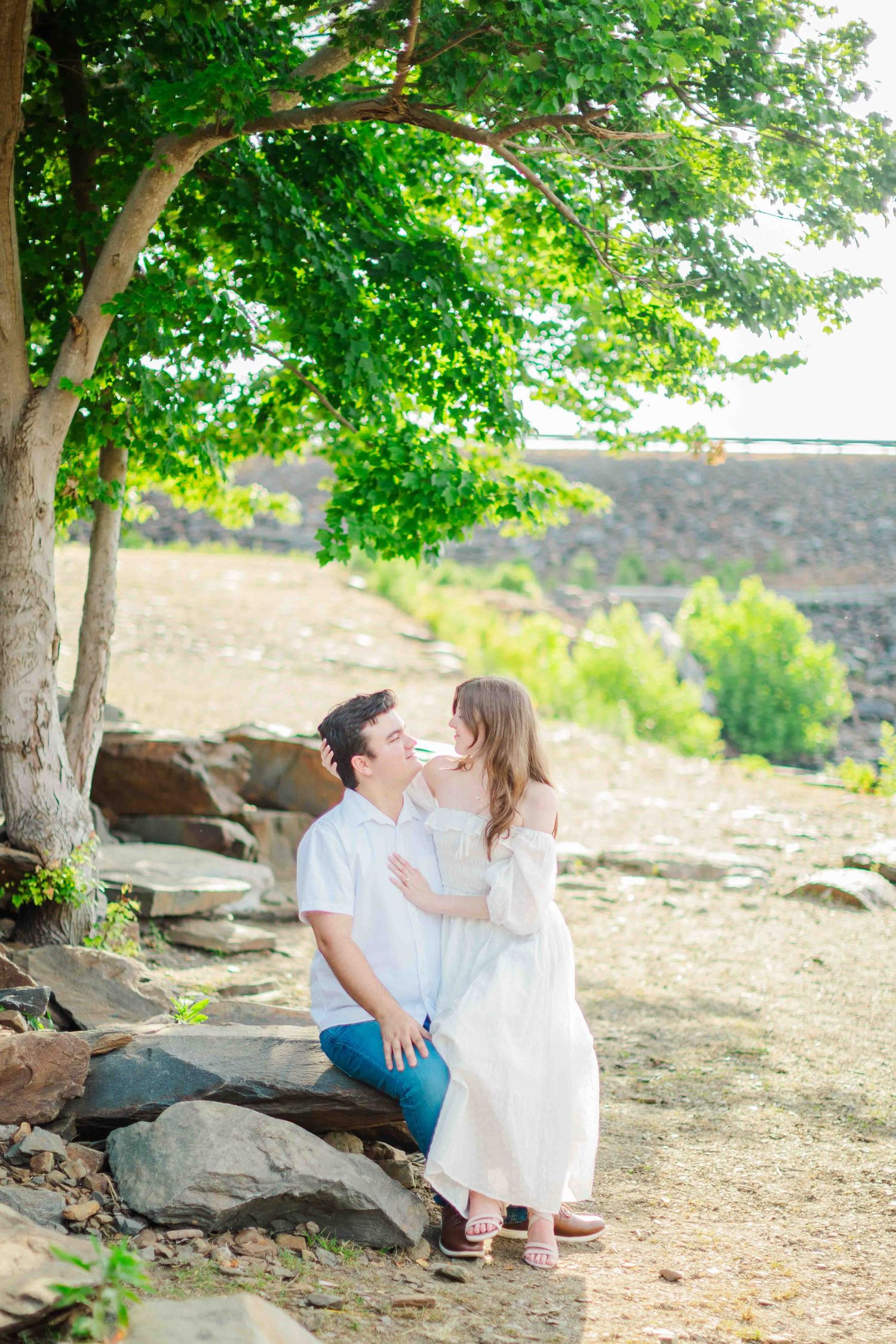 Couple sharing a tender moment under a green tree, woman in white dress, man in white shirt and jeans.