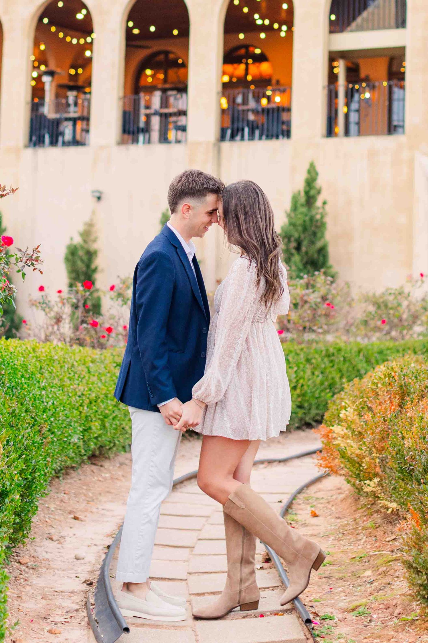 Couple leaning in for a kiss in a garden pathway with string lights and greenery in background.