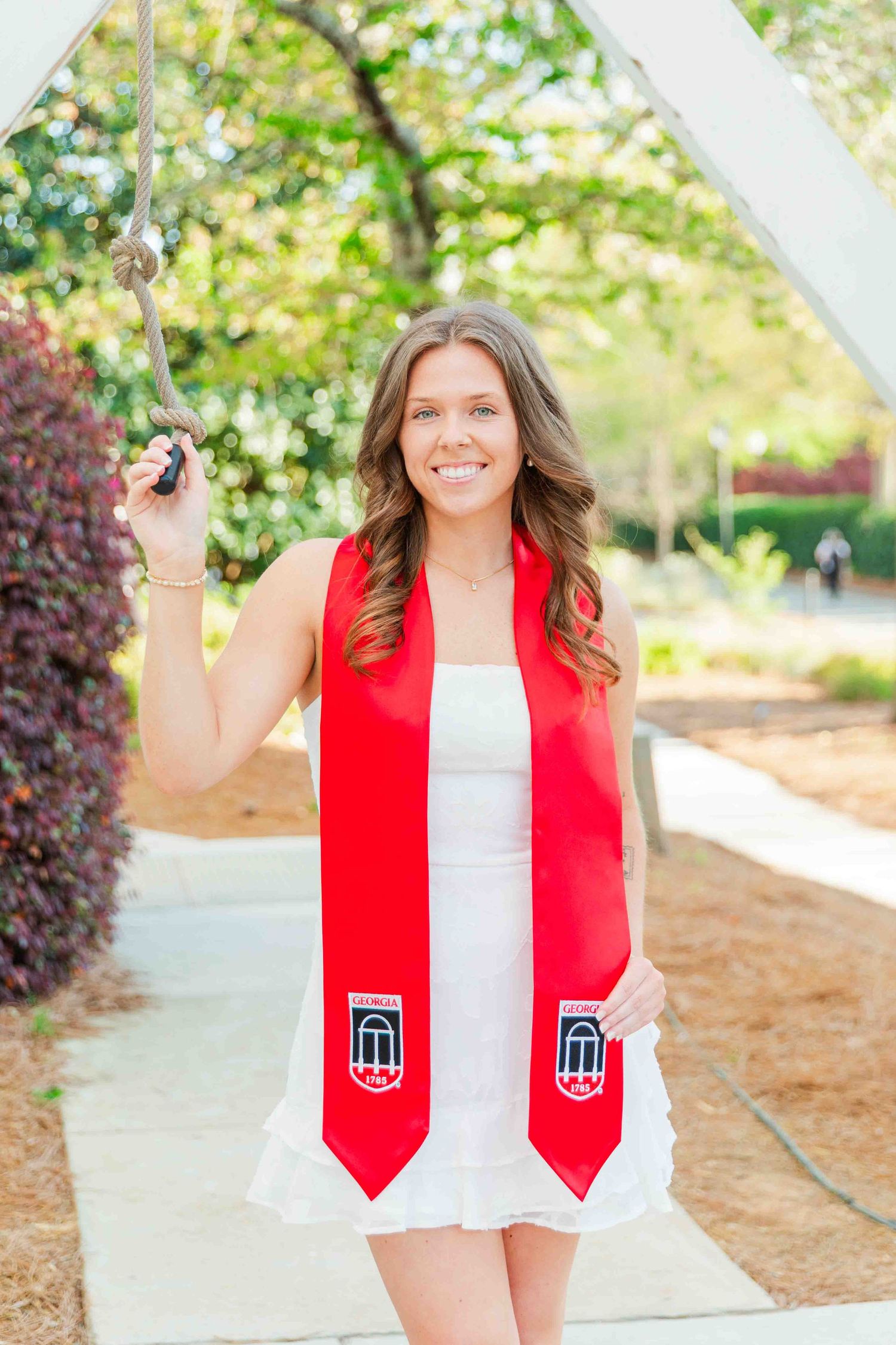 Smiling graduate in white dress and red Georgia sash poses under campus archway.