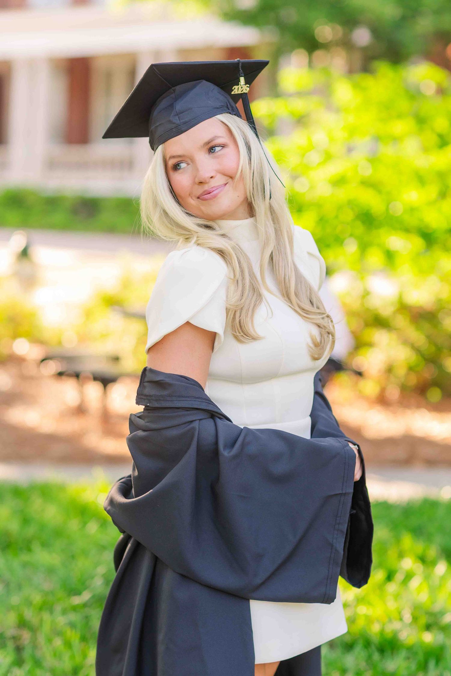 Smiling blonde woman in graduation cap and gown holding diploma outdoors on sunny campus.