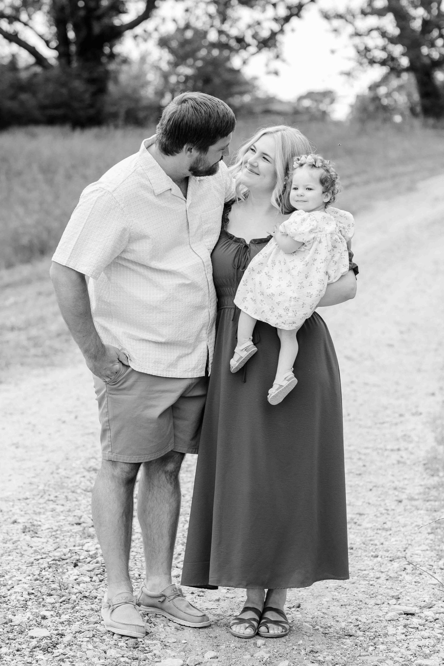 Black and white family portrait of couple with toddler girl on gravel path surrounded by trees.