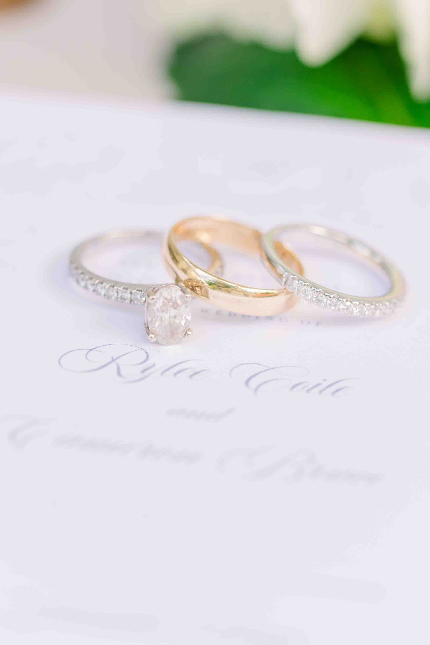 Three wedding rings including a diamond solitaire engagement ring and two gold bands displayed on a white wedding invitation.