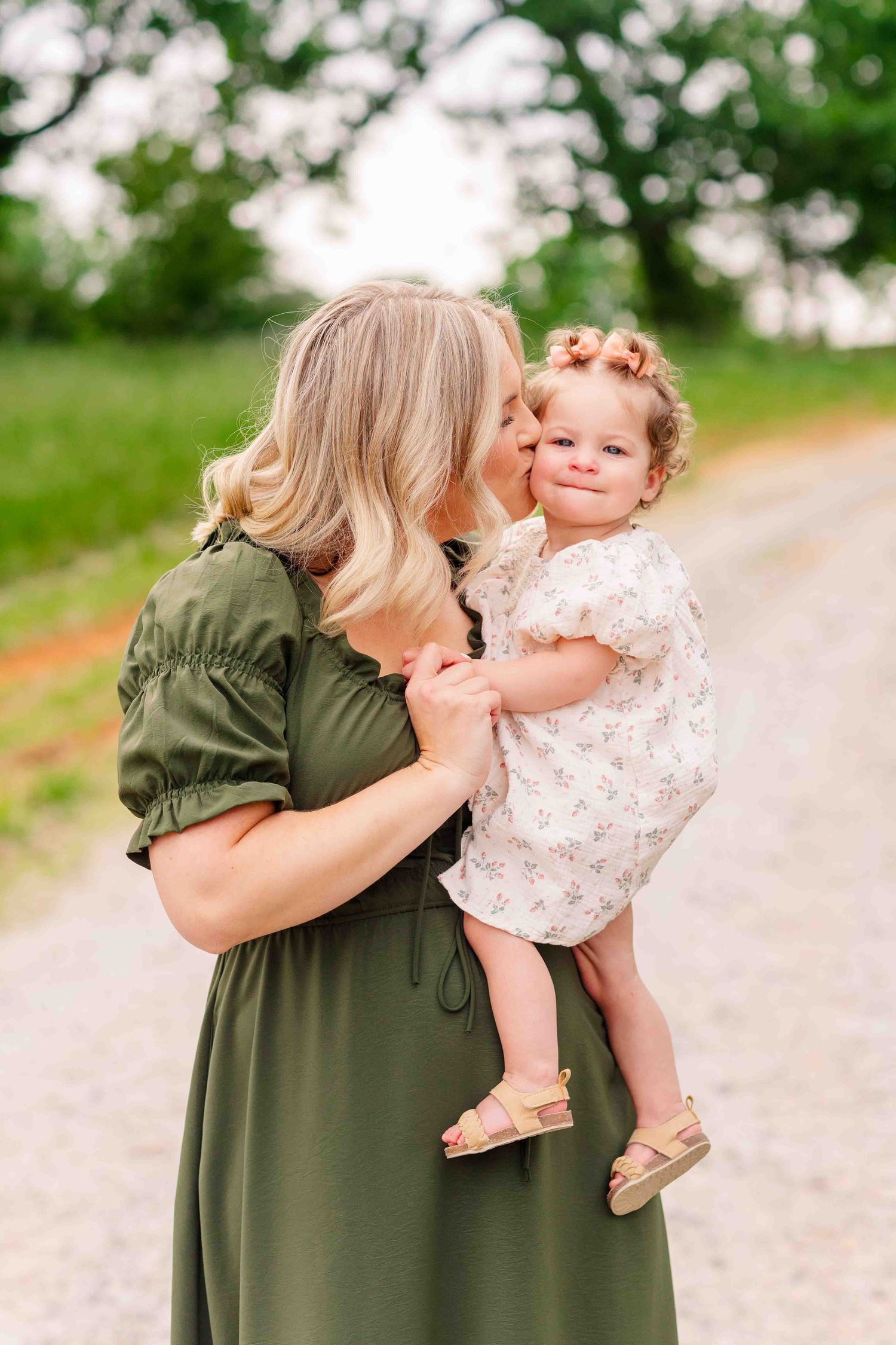 Mother in green dress kissing her curly-haired toddler daughter on a scenic gravel path.