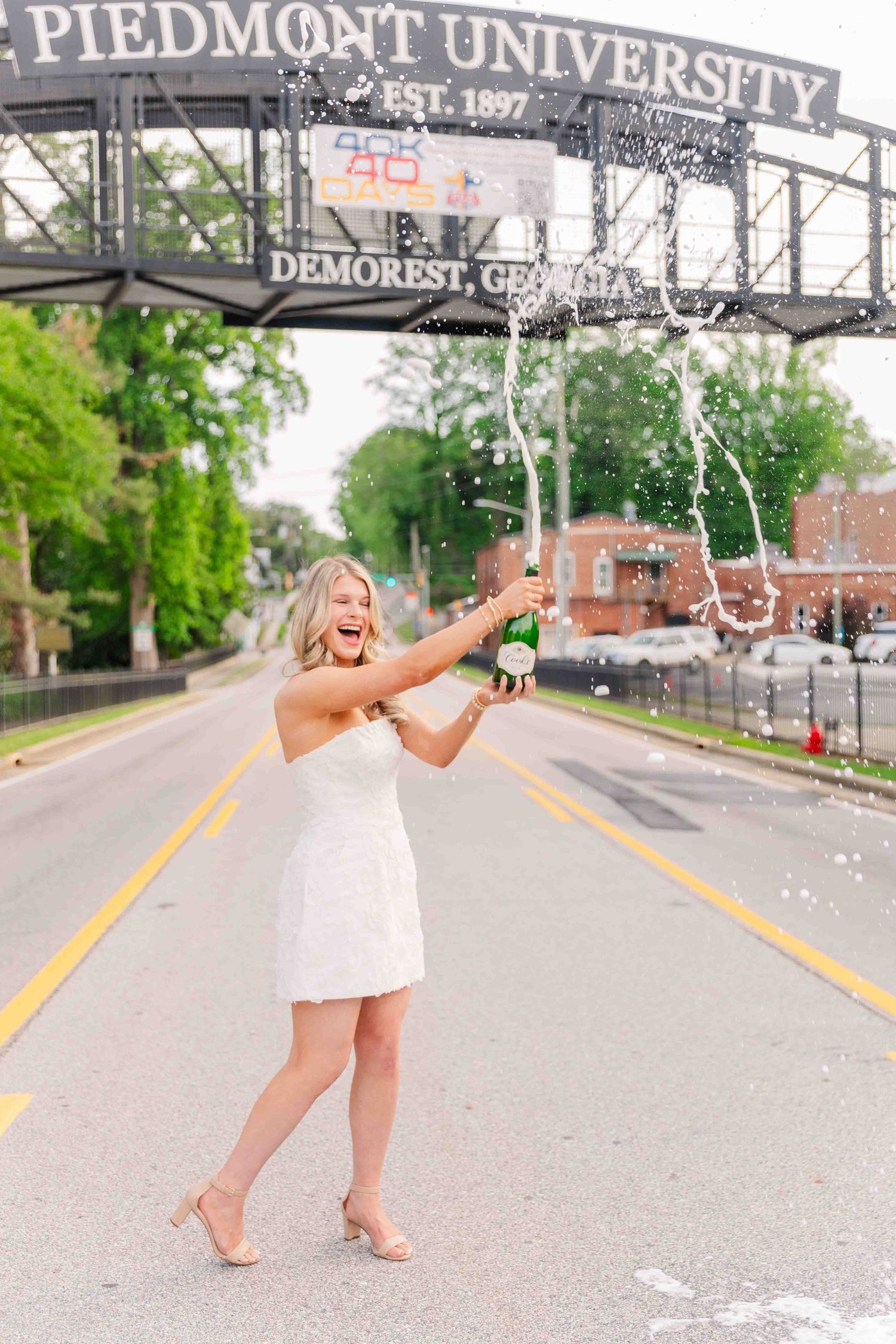 Excited blonde woman popping champagne in front of Piedmont University sign in Demorest Georgia.