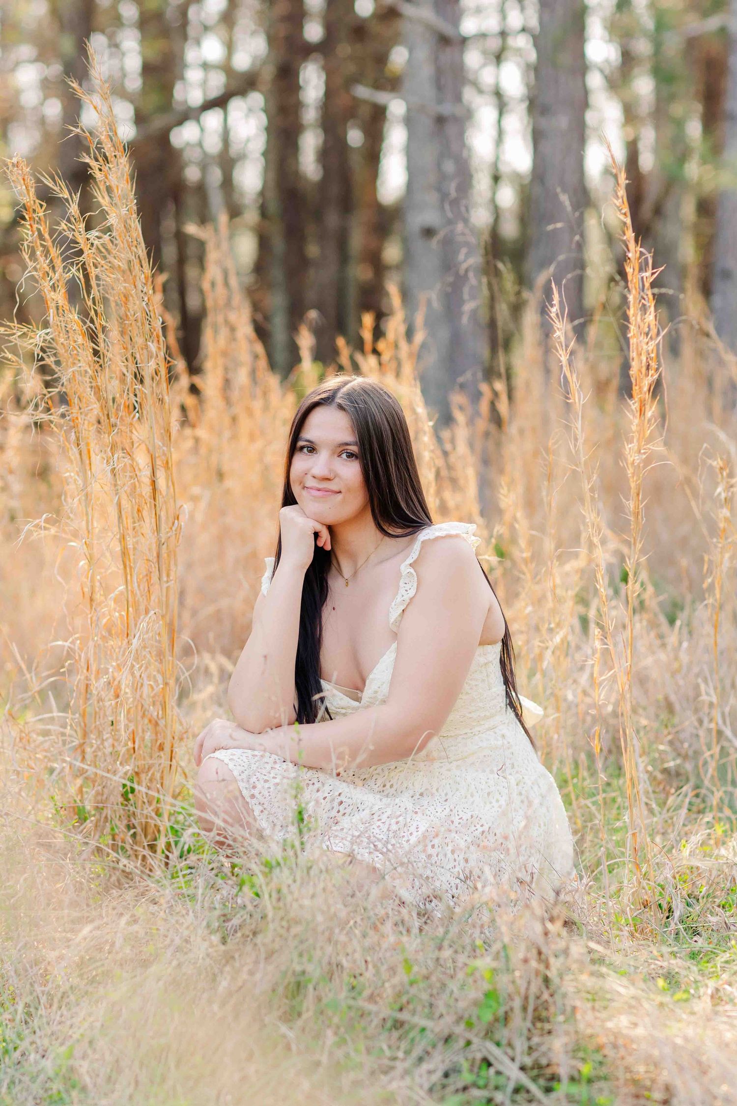 Young brunette woman in lace dress sitting among tall golden grasses in a pine forest.