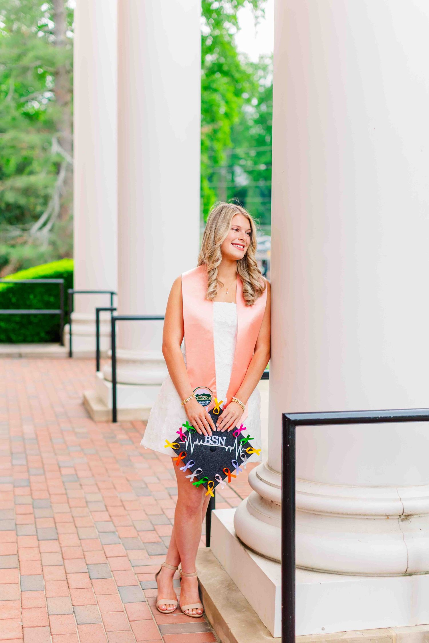 Smiling blonde graduate in pink stole holding BSN graduation cap between white columns on campus.