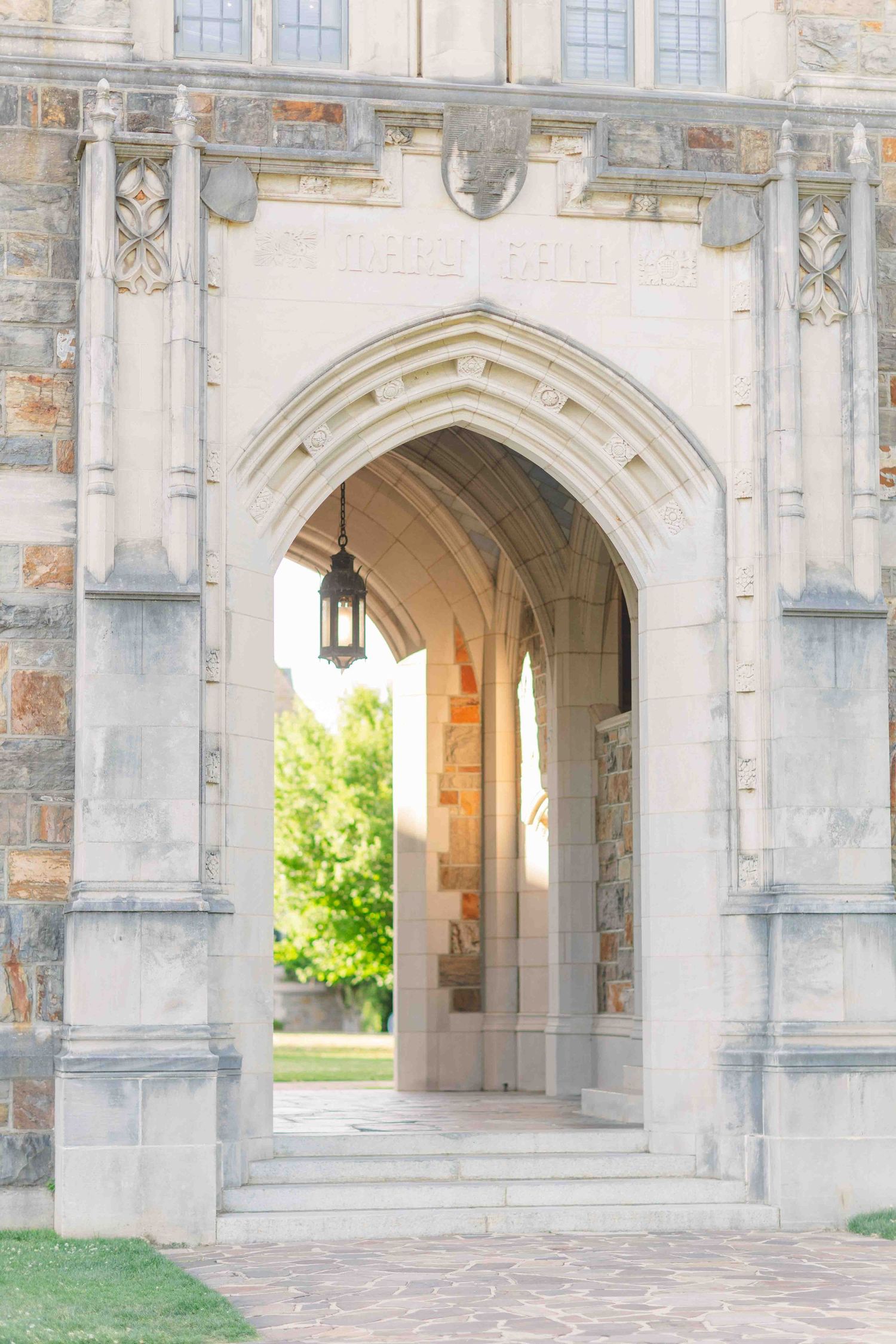 Gothic stone archway with hanging lantern frames a sunlit courtyard at a university campus.