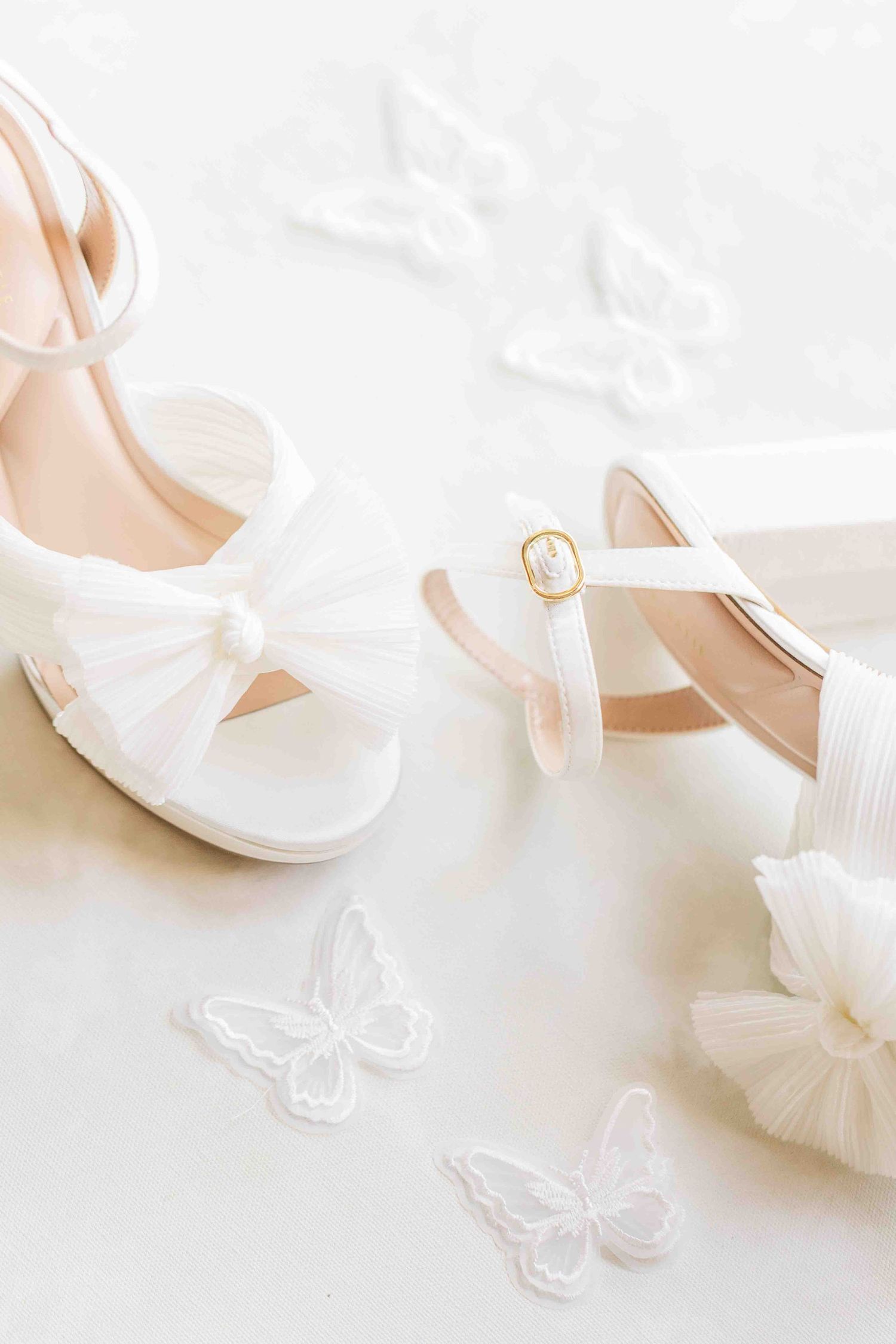 White bridal heels with tulle bow detail surrounded by delicate white butterfly embellishments on a white background.