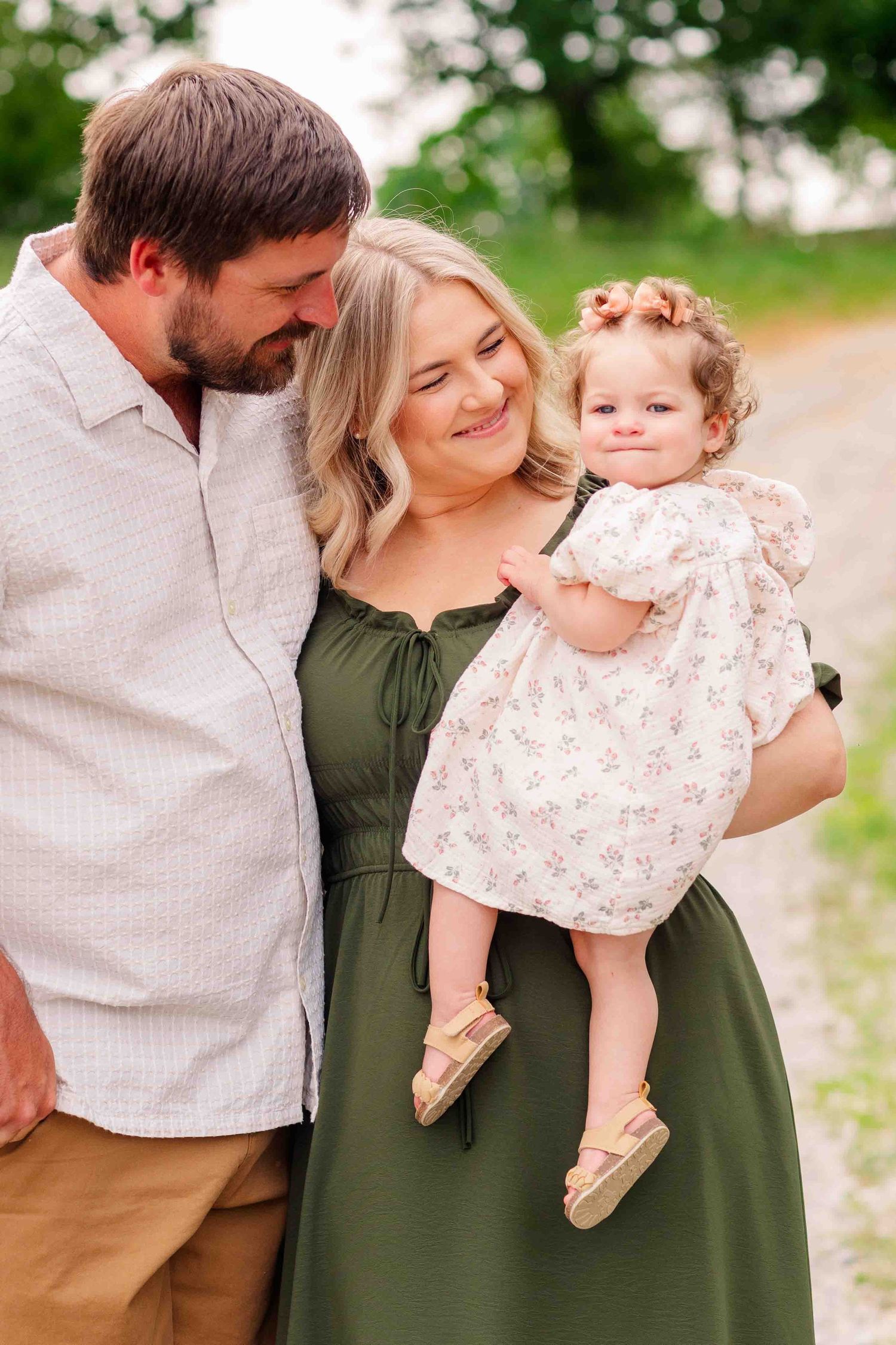 Smiling couple in green and white outfits holding their baby girl in a floral dress outdoors.