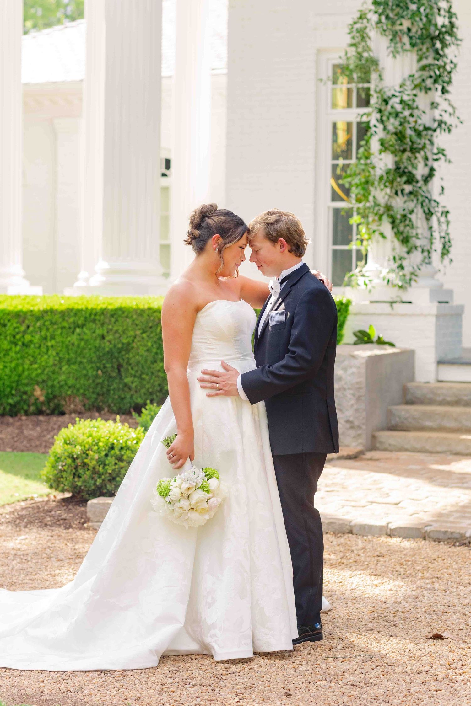 Bride and groom lean in for a kiss outside a grand white columned estate, bride holding a white floral bouquet.