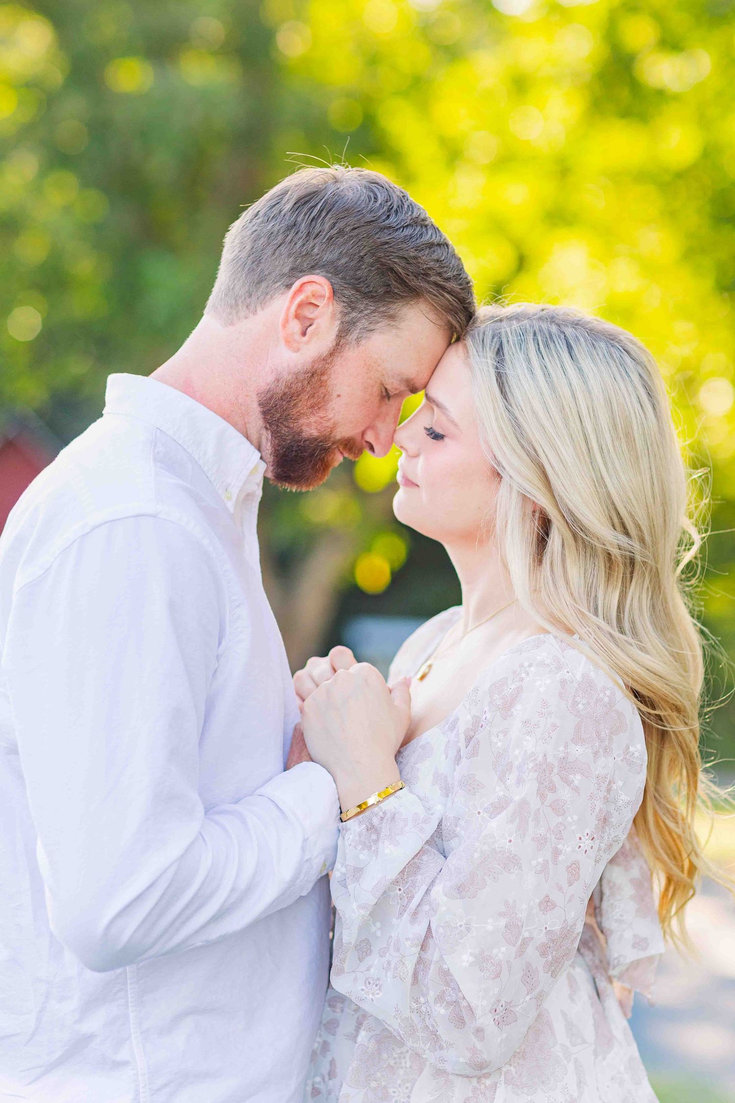 Engaged couple holding hands and touching foreheads tenderly in warm golden sunlight outdoors.