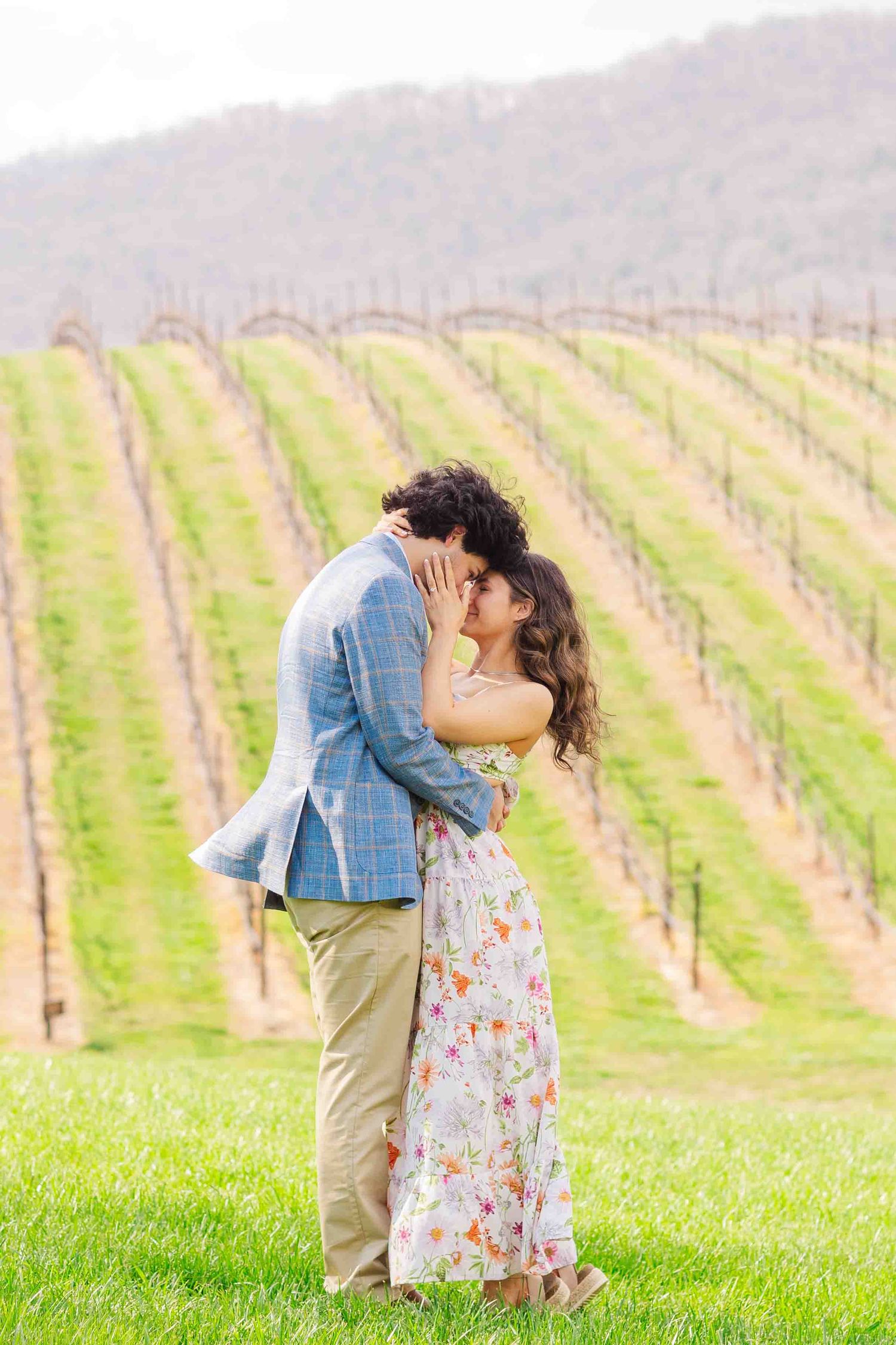 Couple sharing a passionate kiss on lush green grass with scenic vineyard rows and mountain backdrop.