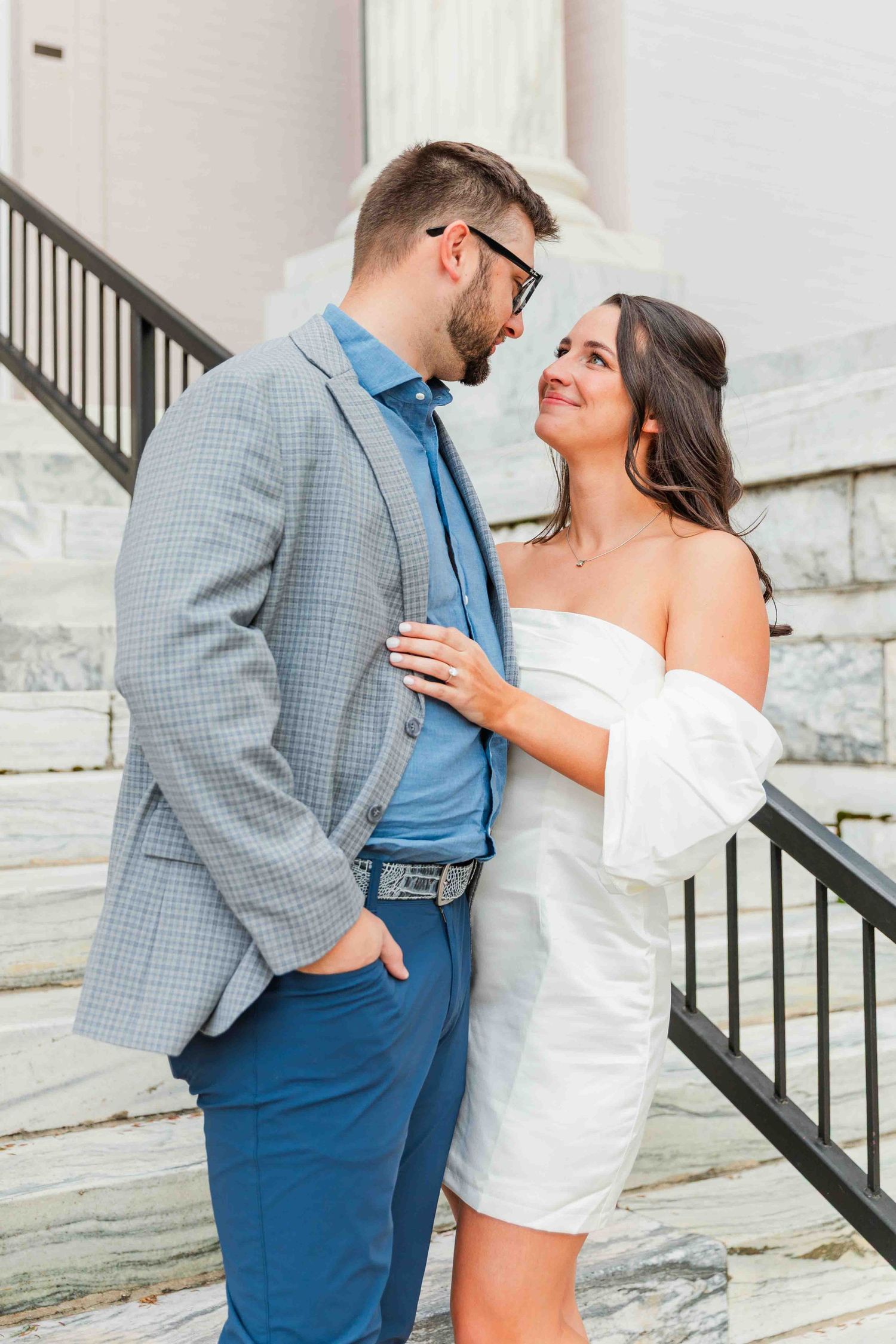 Couple gazing at each other on marble steps, man in gray blazer and woman in white off-shoulder dress.