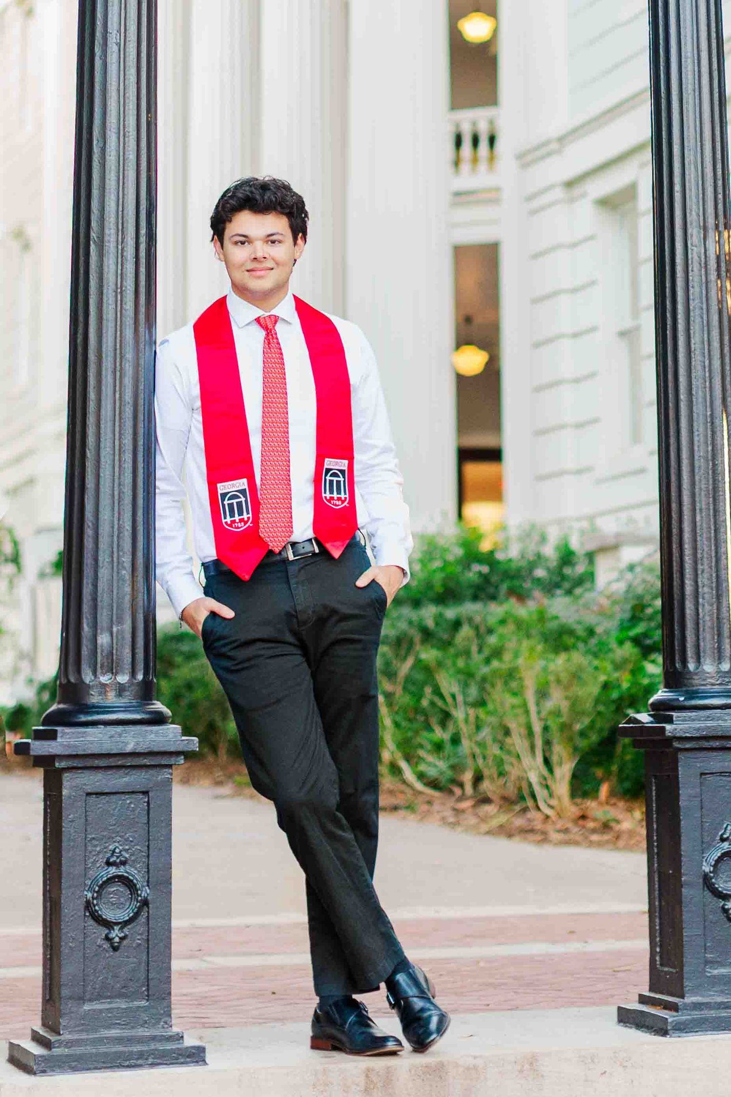 Young man in white shirt, red tie, and red graduation stole leaning against iron poles outside a classical building.