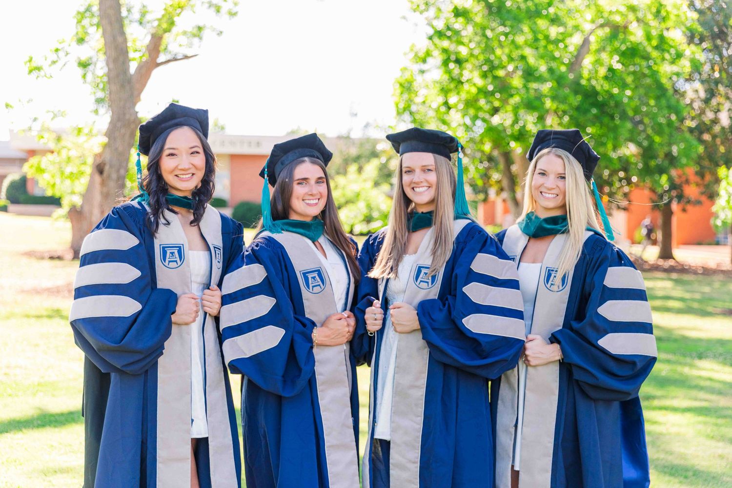 Four smiling doctoral graduates in navy regalia pose together on sunny campus grounds.