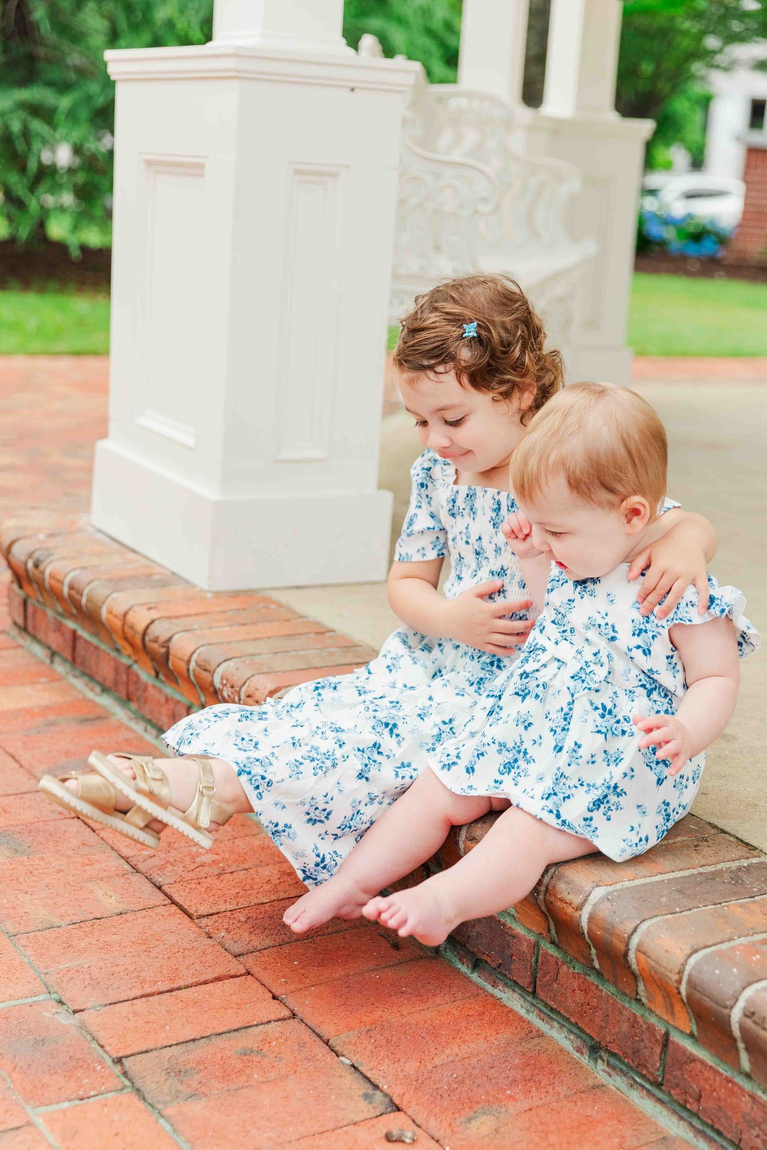 Two young sisters in matching blue floral dresses sitting together on brick steps.