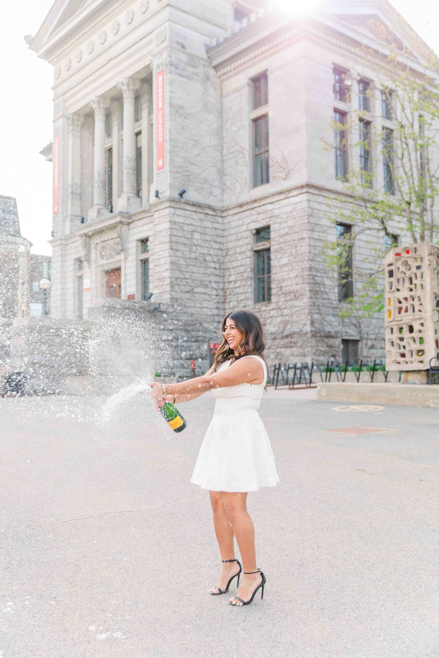 Woman in white dress popping champagne bottle outside historic stone building, celebrating in strappy heels.