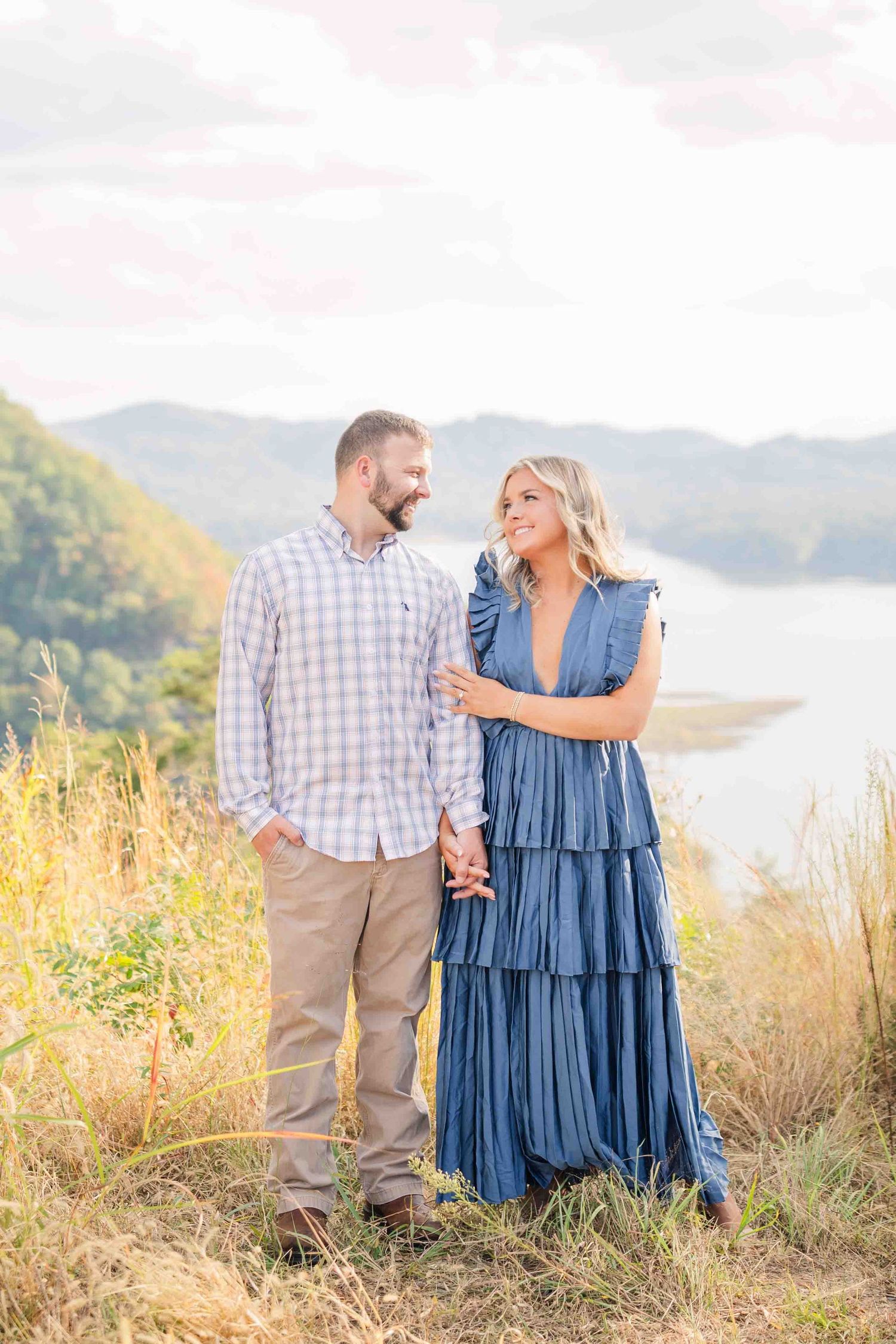 Couple holding hands on a scenic overlook with mountain lake views, woman in blue tiered dress.