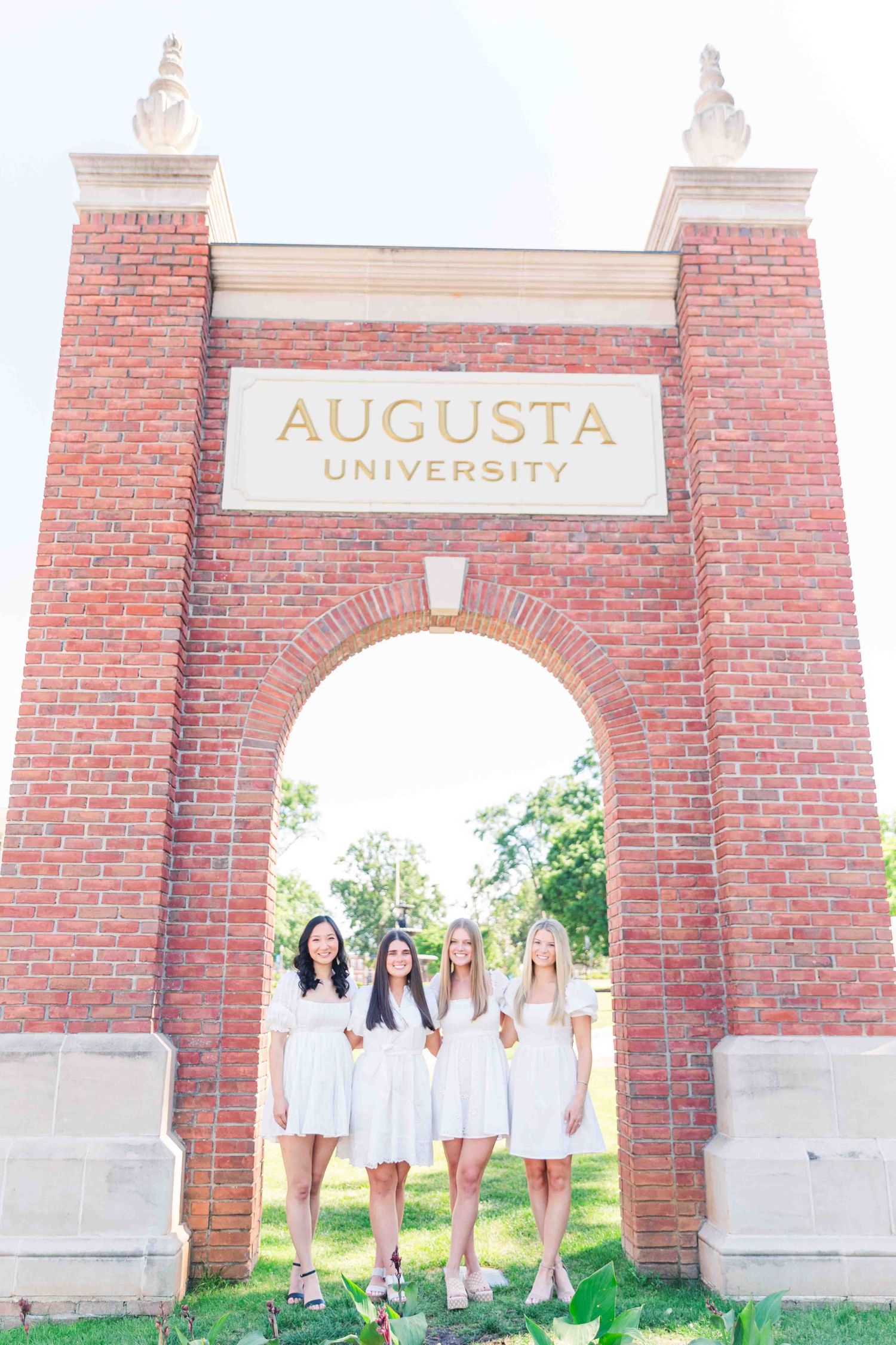 Four women in white dresses pose under Augusta University brick arch entrance.