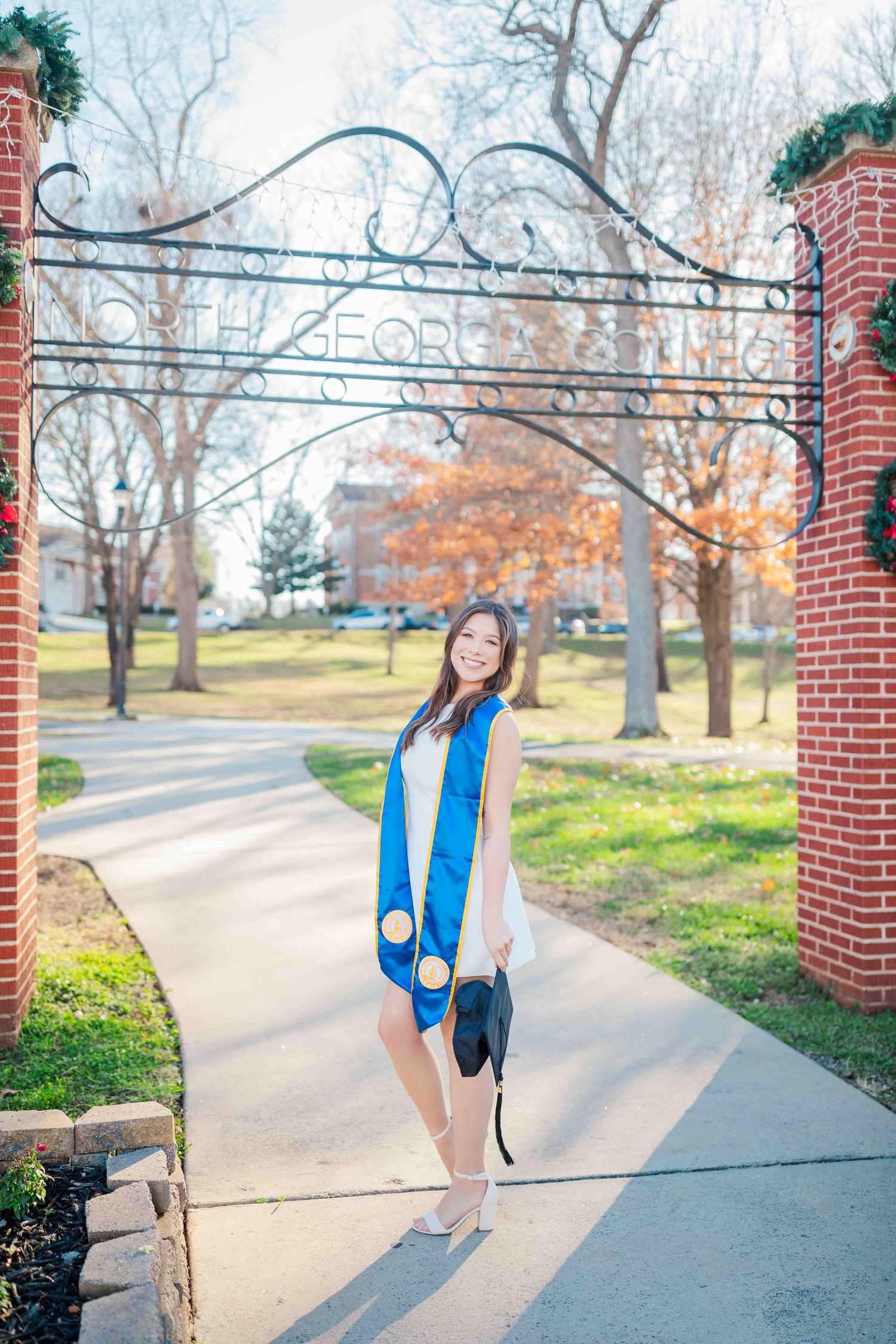 Young woman in blue graduation stole holding cap, posing at North Georgia College entrance gate on a sunny day.