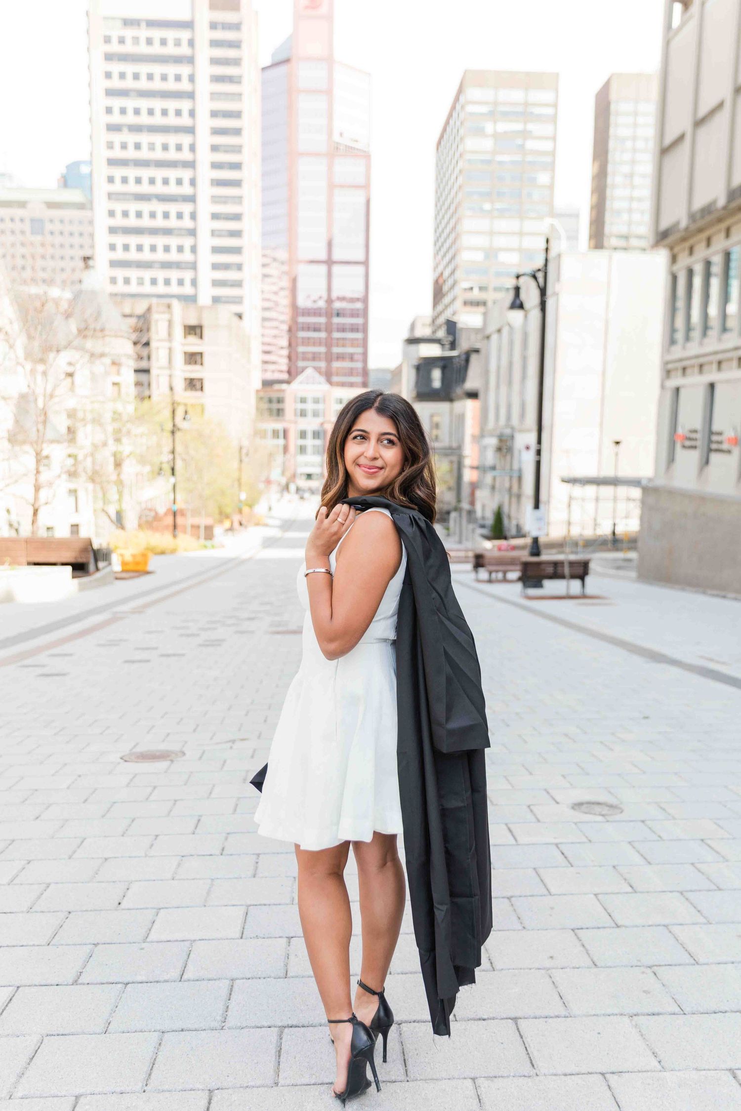 Young woman in white dress holding black graduation gown, posing on urban plaza with city skyscrapers in background.