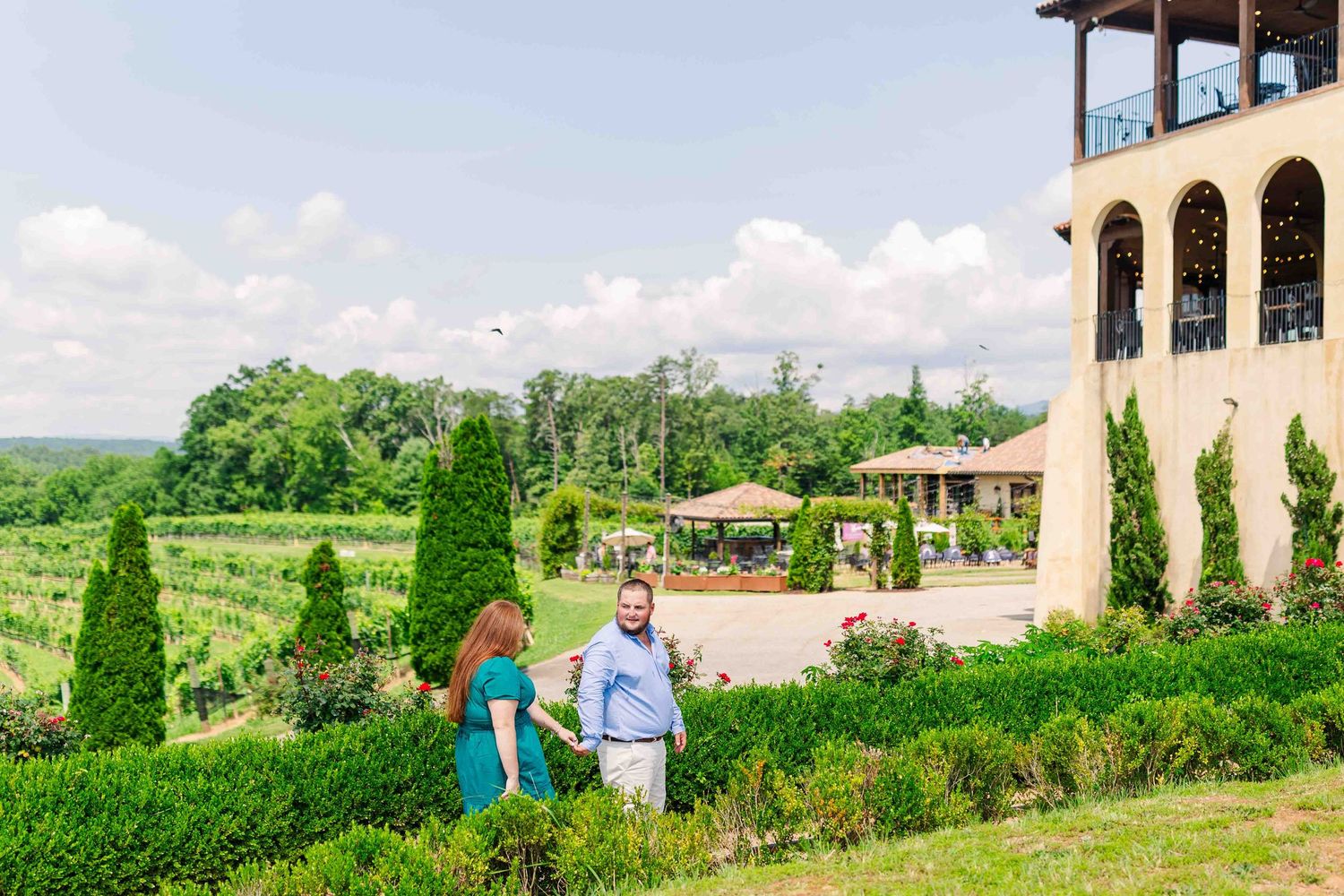 Couple walking hand in hand through manicured winery gardens with rolling vineyard and elegant estate in background.