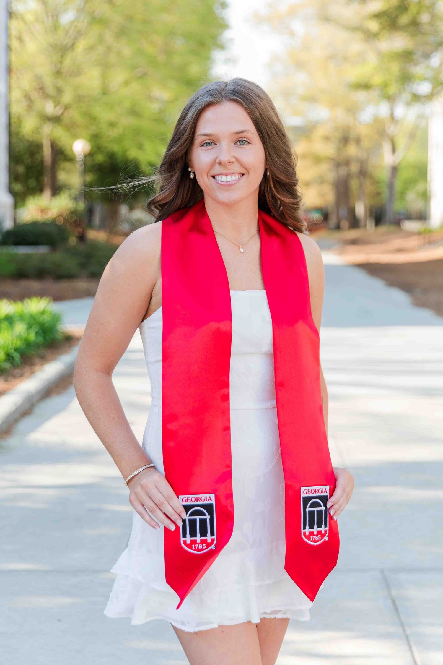 Smiling graduate wearing red Georgia university sash poses on sunny campus walkway.