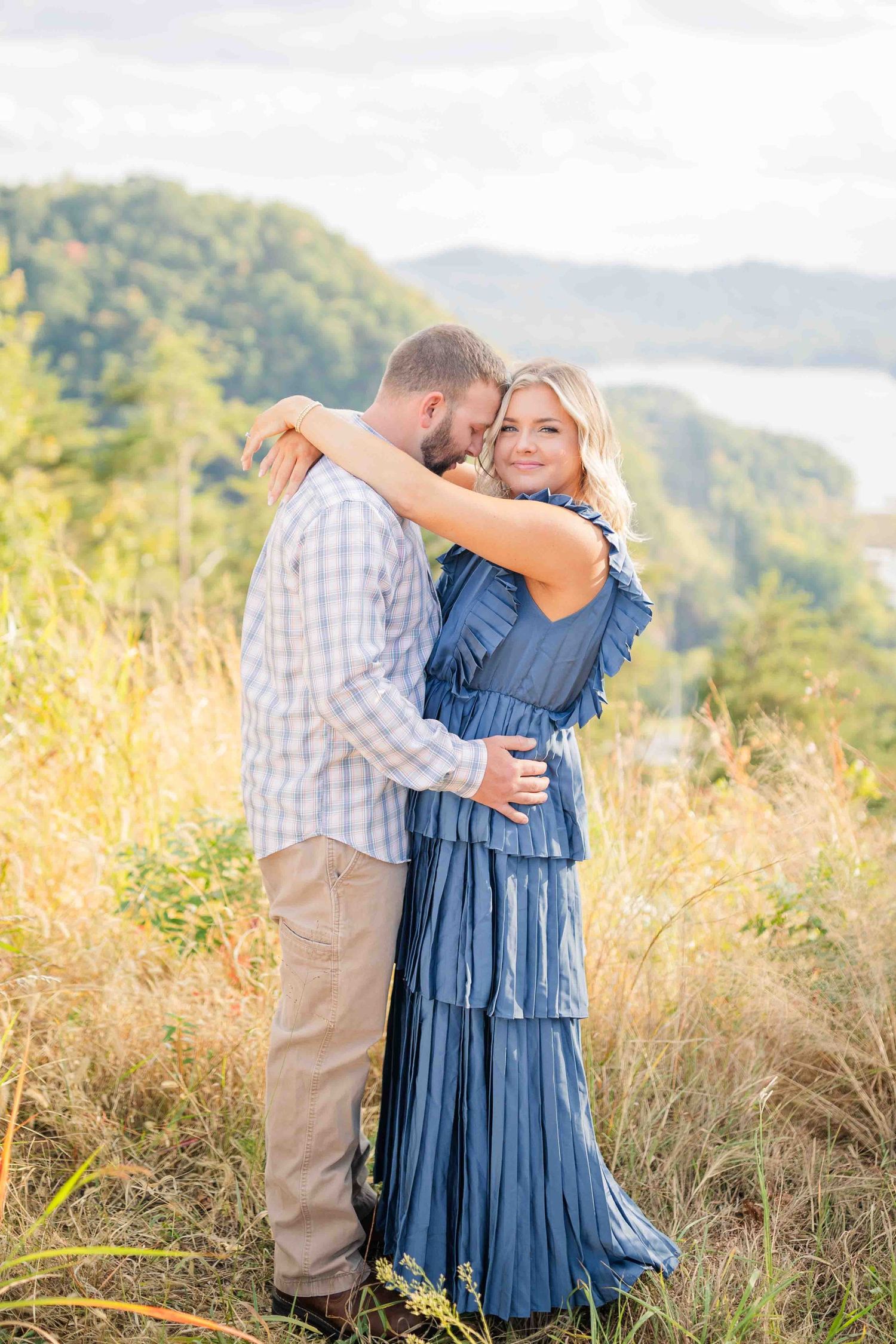 Couple embracing on a scenic hilltop, woman in blue maxi dress smiling, lush green mountains and lake in background.