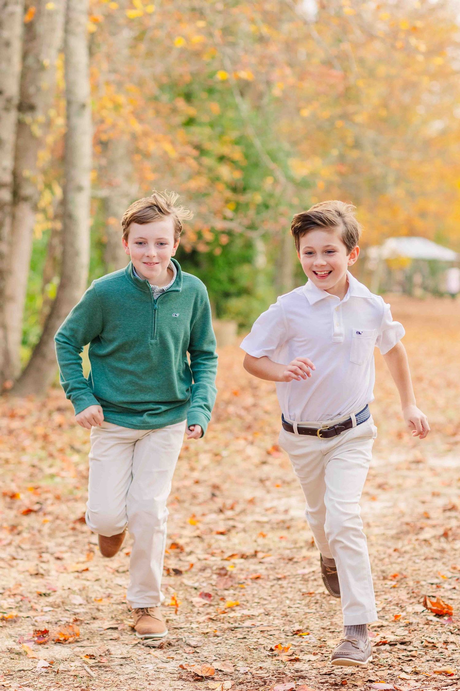 Two boys in green and white shirts running and laughing along a leaf-covered autumn path.