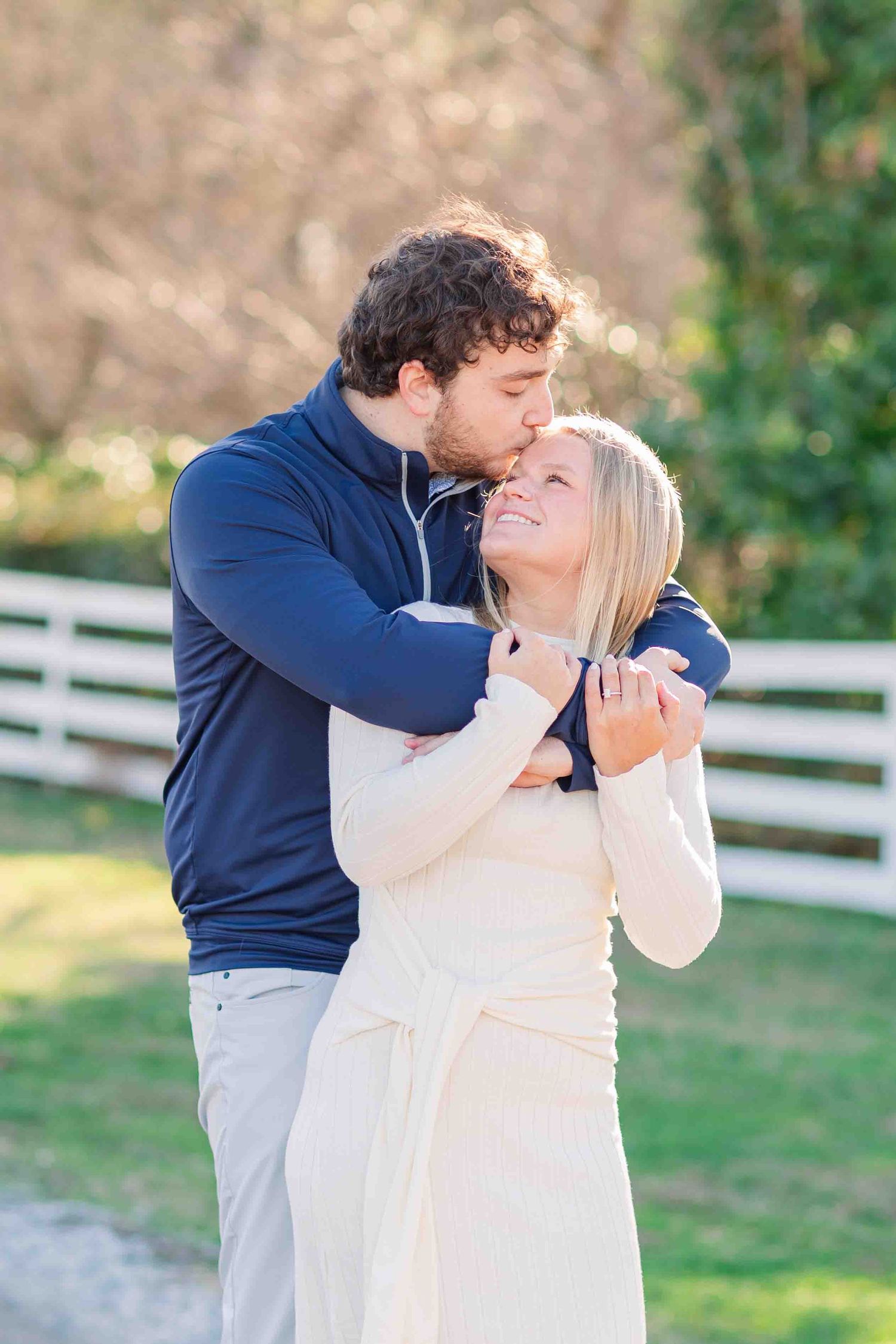 Man in navy pullover kissing woman on forehead while embracing near white fence in golden light.